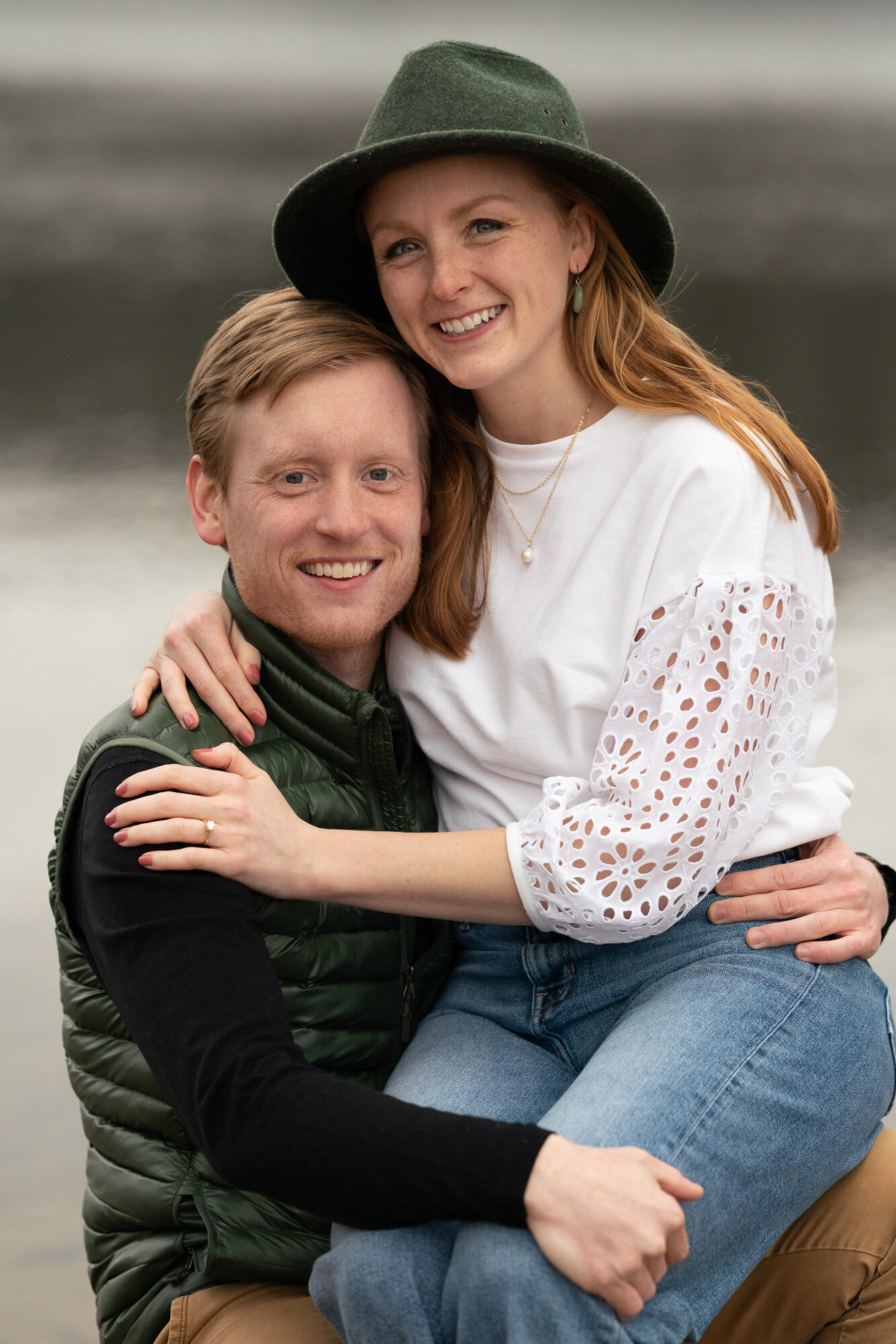 Woman wearing white sweater and green hat smiles while sitting on her fiancee's lap.