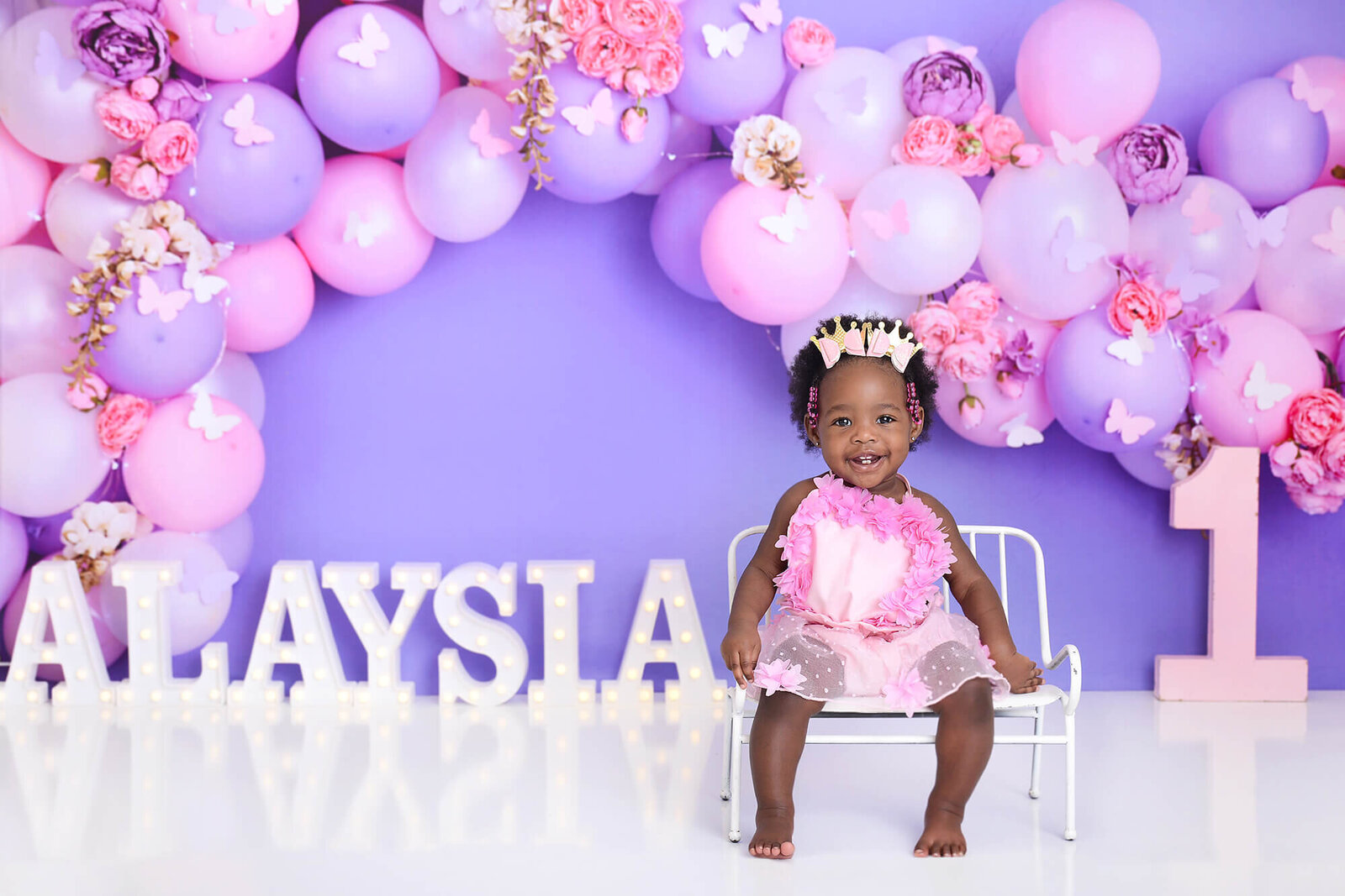 girl smiles at camera at her first birthday photoshoot in houston, in front of a purple butterfly background