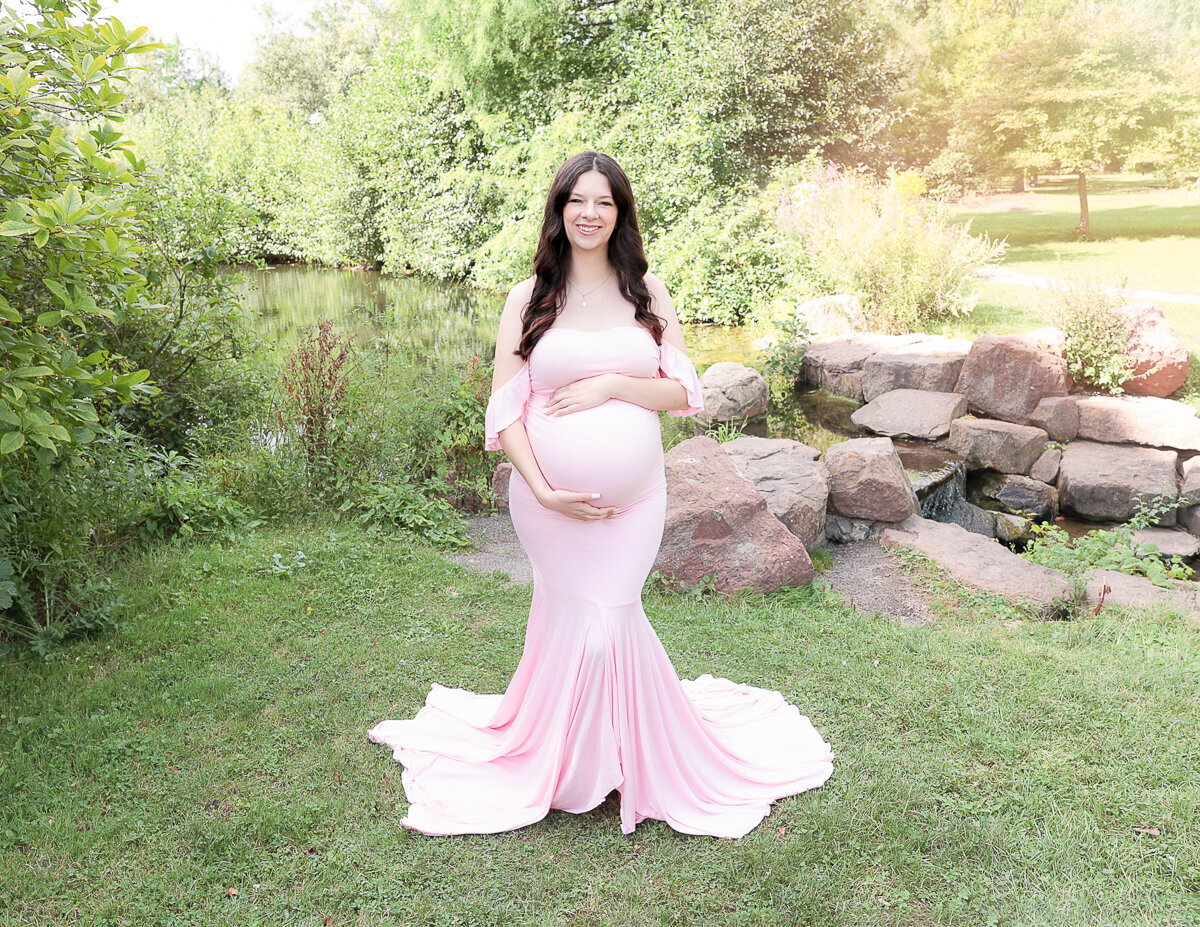 Expecting mother standing in an outdoor setting, gently cradling her baby bump during a maternity  photography session in Rochester, NY. The soft natural light highlights her glowing skin and the beauty of motherhood.