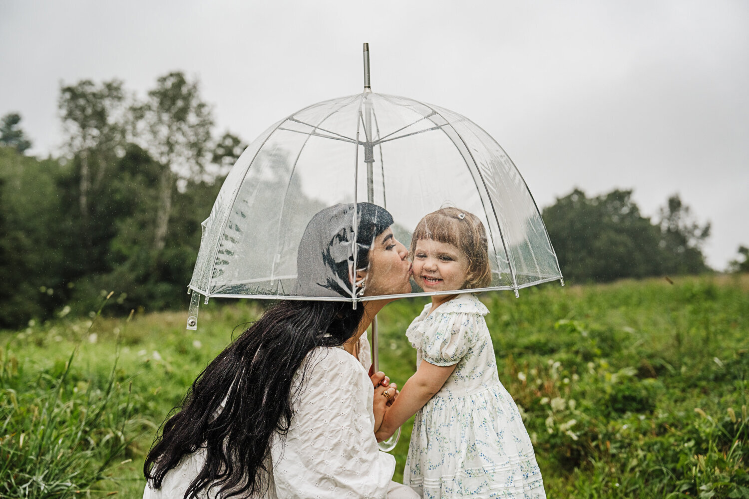 mom kisses little girl under umbrella in a rainy field
