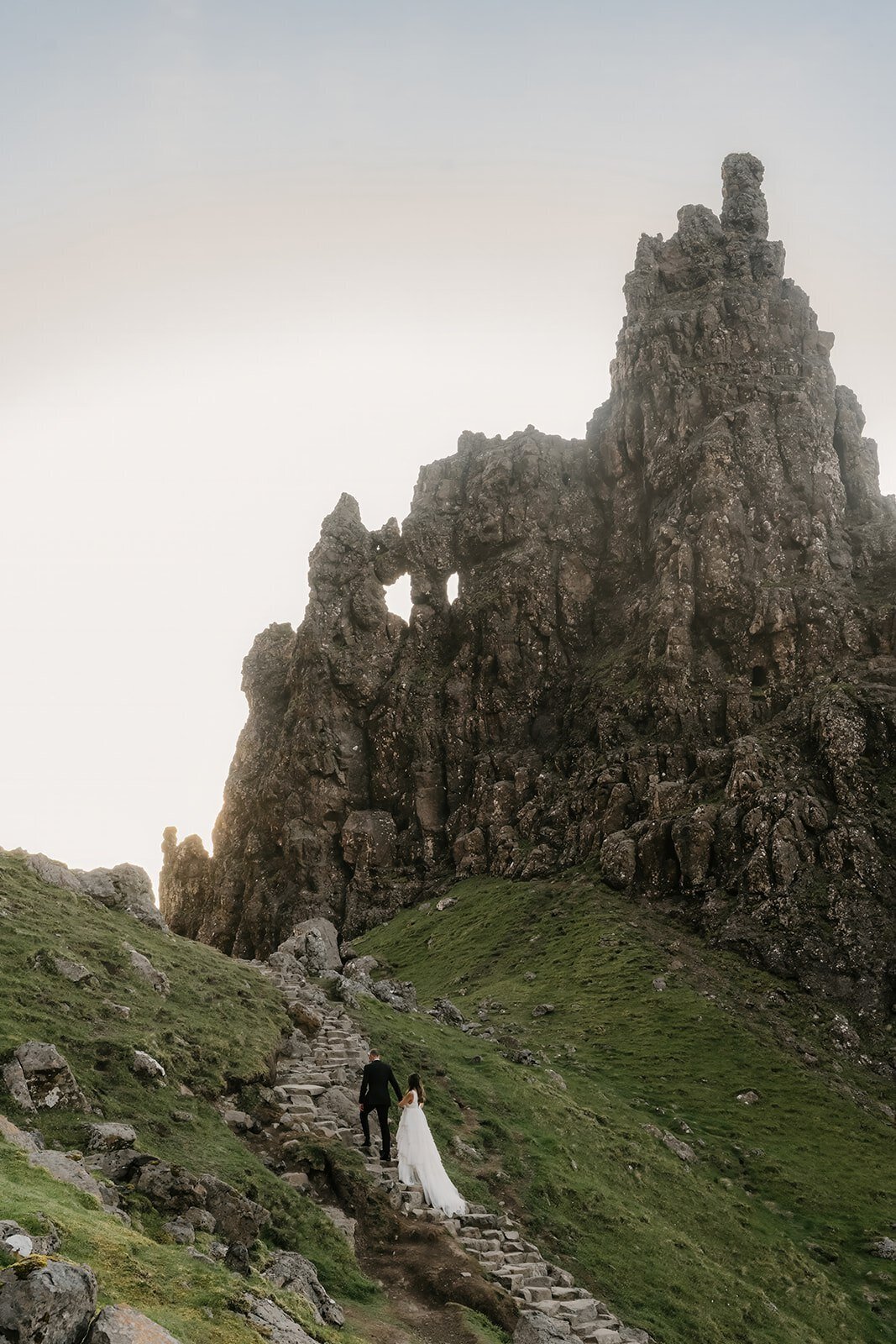 A bride and groom walk up stone steps toward a rocky mountain under a bright sky—a scene perfect for a Switzerland photo retreat in the Alps for adventurous photographers.