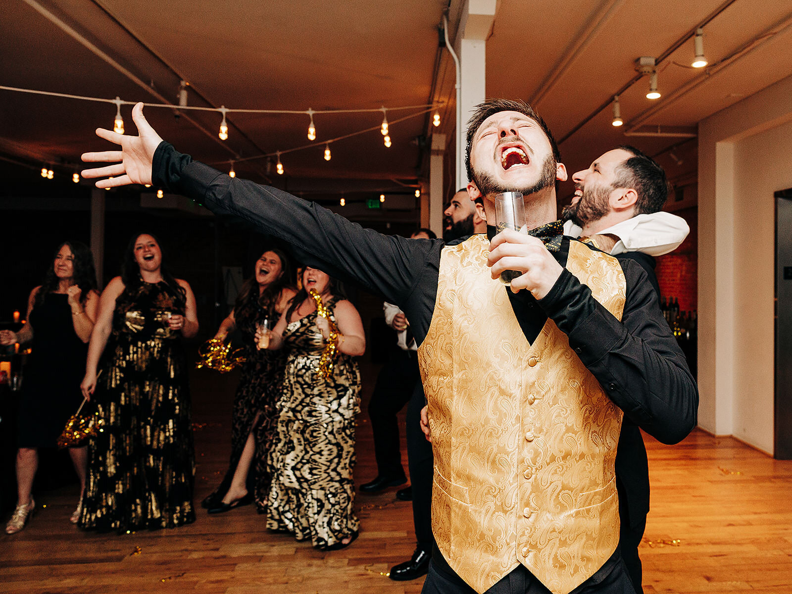 A man in a gold vest sings passionately with a drink in hand at a lively party. Captured by a Denver Wedding Photographer, guests in formal attire laugh and cheer under string lights in this festive indoor setting.