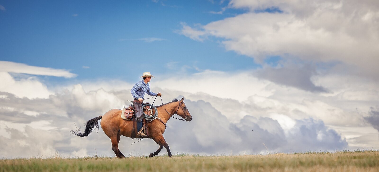 Senior boy  in cowboy gear galloping on a horse with a blue sky and clouds in the background