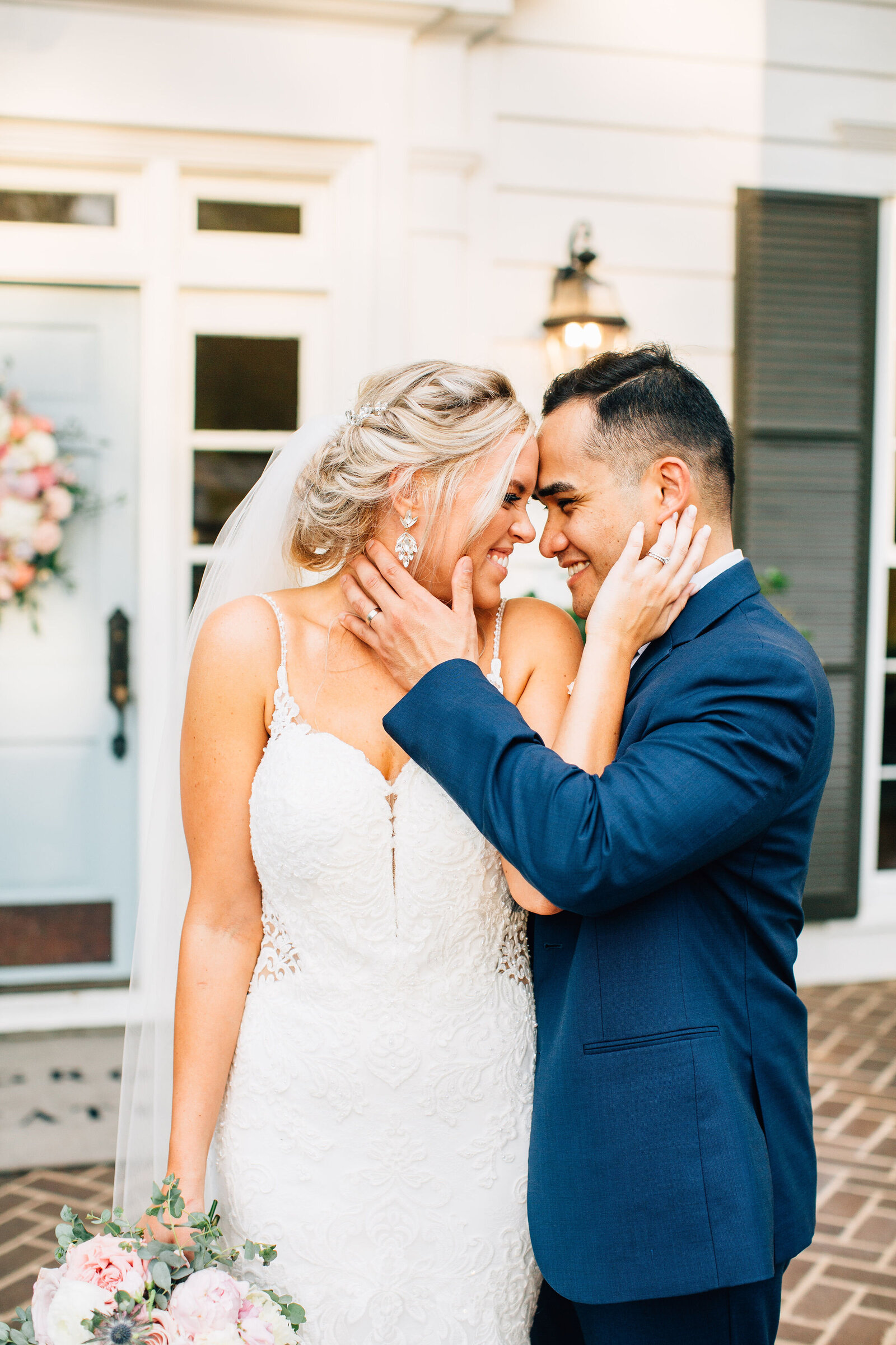 bride with blond hair, wearing a white wedding dress and her groom with brown hair wearing  a blue suit, stand outside a house touching each others cheeks and smiling