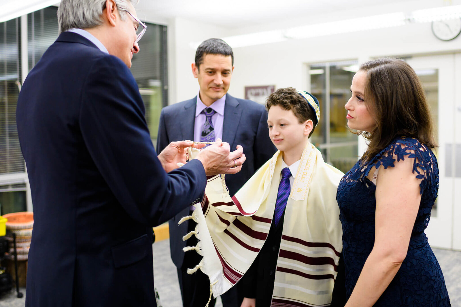 A teen boy in a Kippah and Tallit speaks with a Rabbi about the cords on his tallit during his bar mitzvah with mom and dad