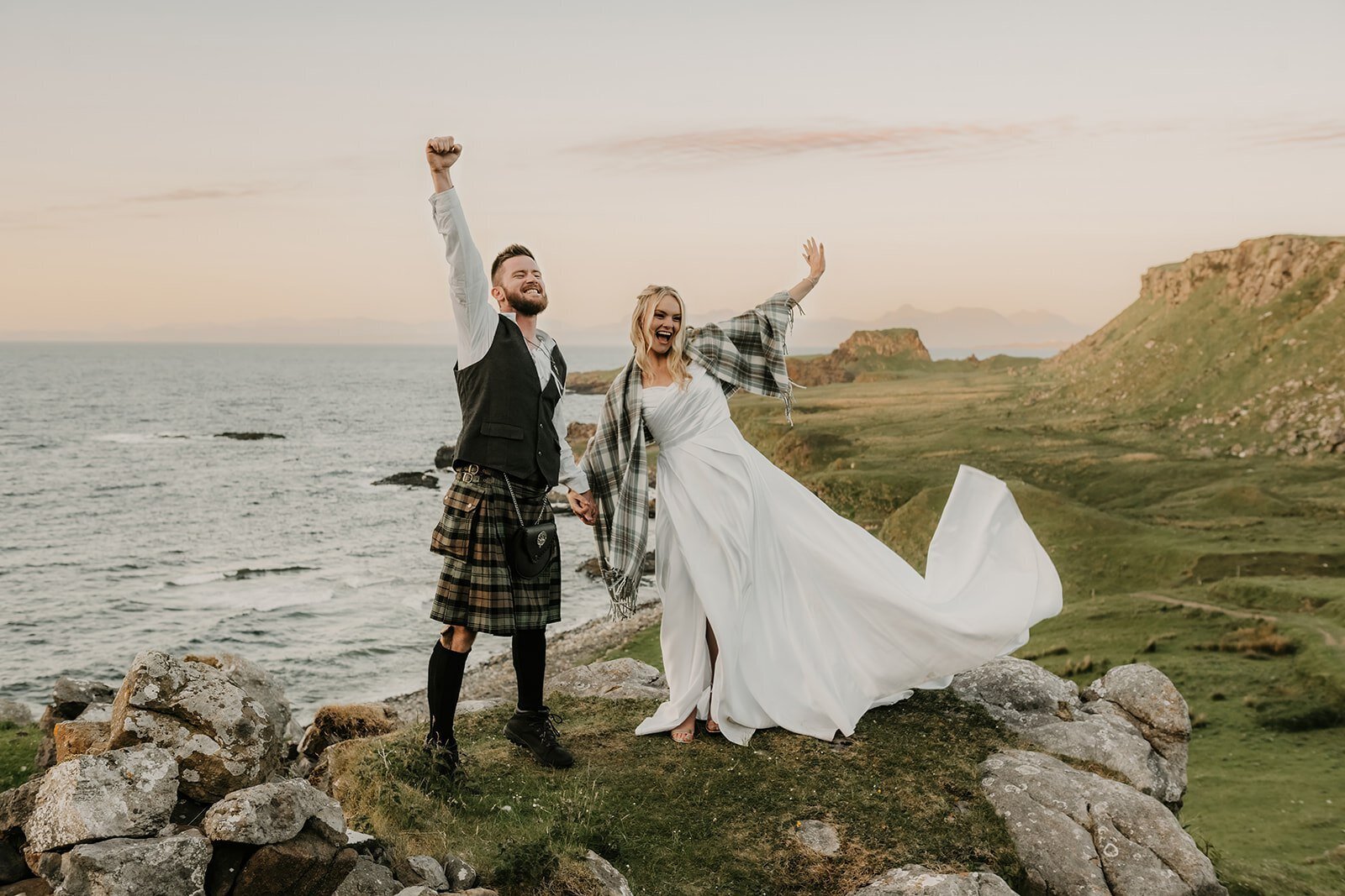 A joyful bride and groom in wedding attire celebrate on rocky cliffs by the sea at sunset, channeling the spirit of an Oregon coast photography retreat for wild-hearted artists.