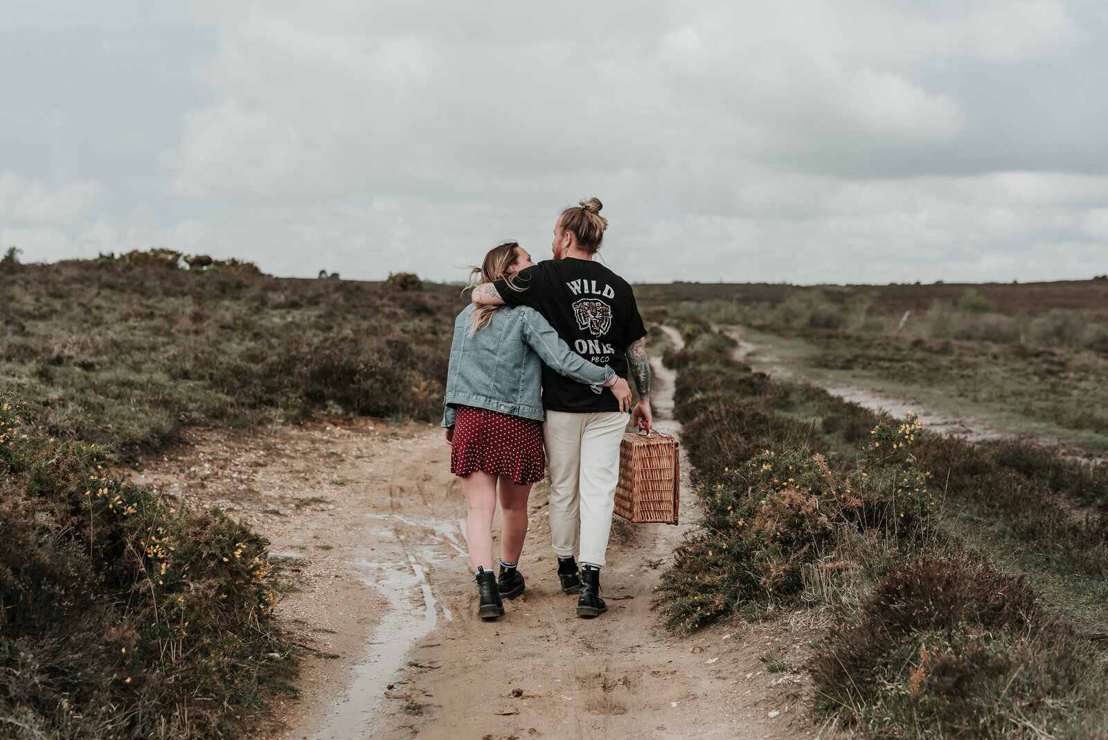 Couple holding a picnic basket walk away hugging eachother along a muddy pathway in the moorlands of New Forest