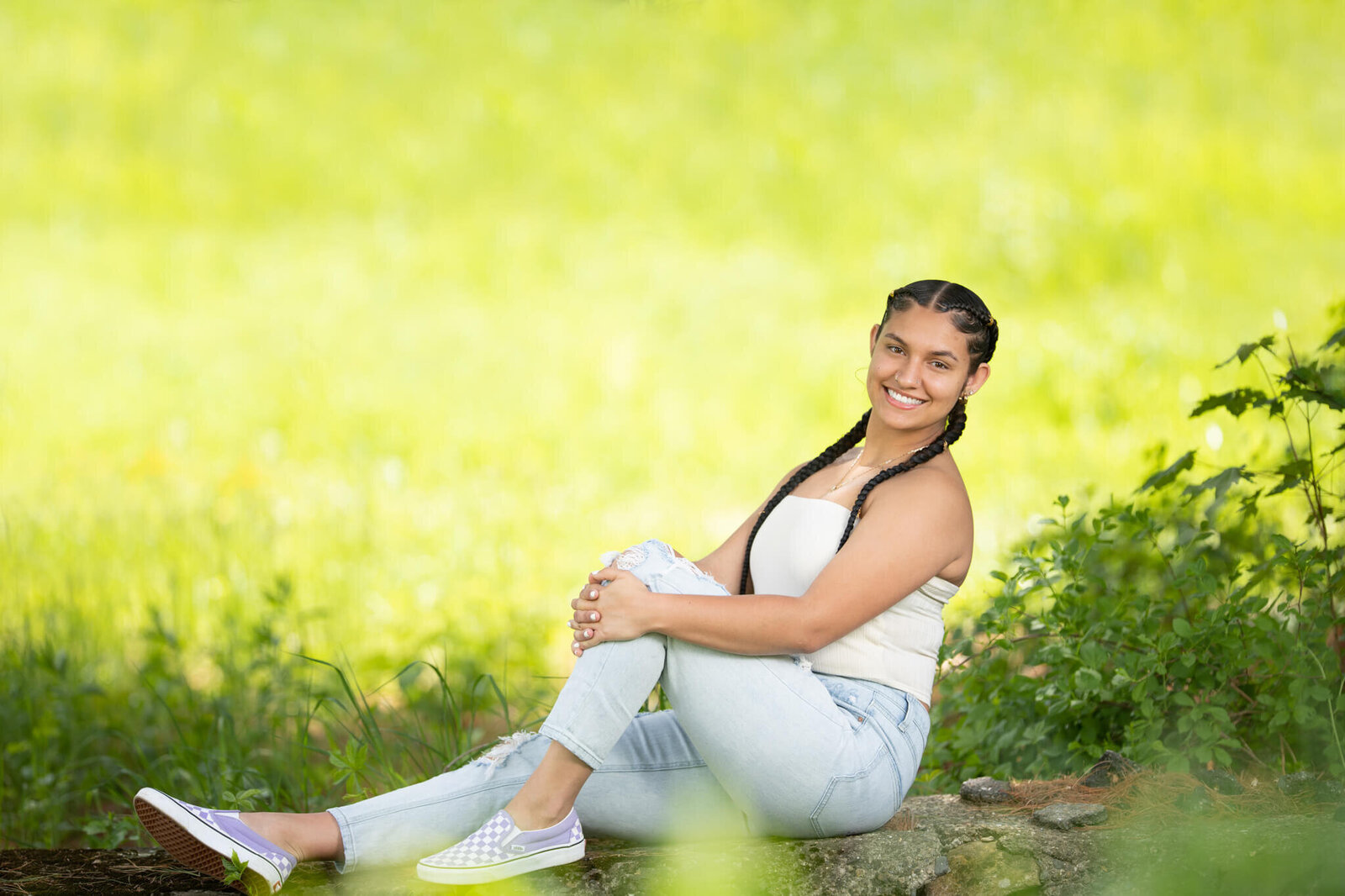 High school senior portrait photography in Clinton MA  of female in a white top smiling at the camera with a green and yellow background sitting on the rock wall