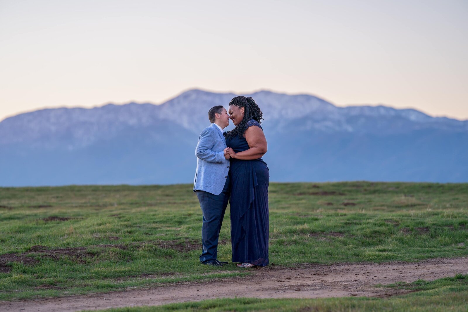 Black skinned woman wearing blue dress holding both hands of woman in blue pants and lightblue jacket kissing in front of mountain sunset.