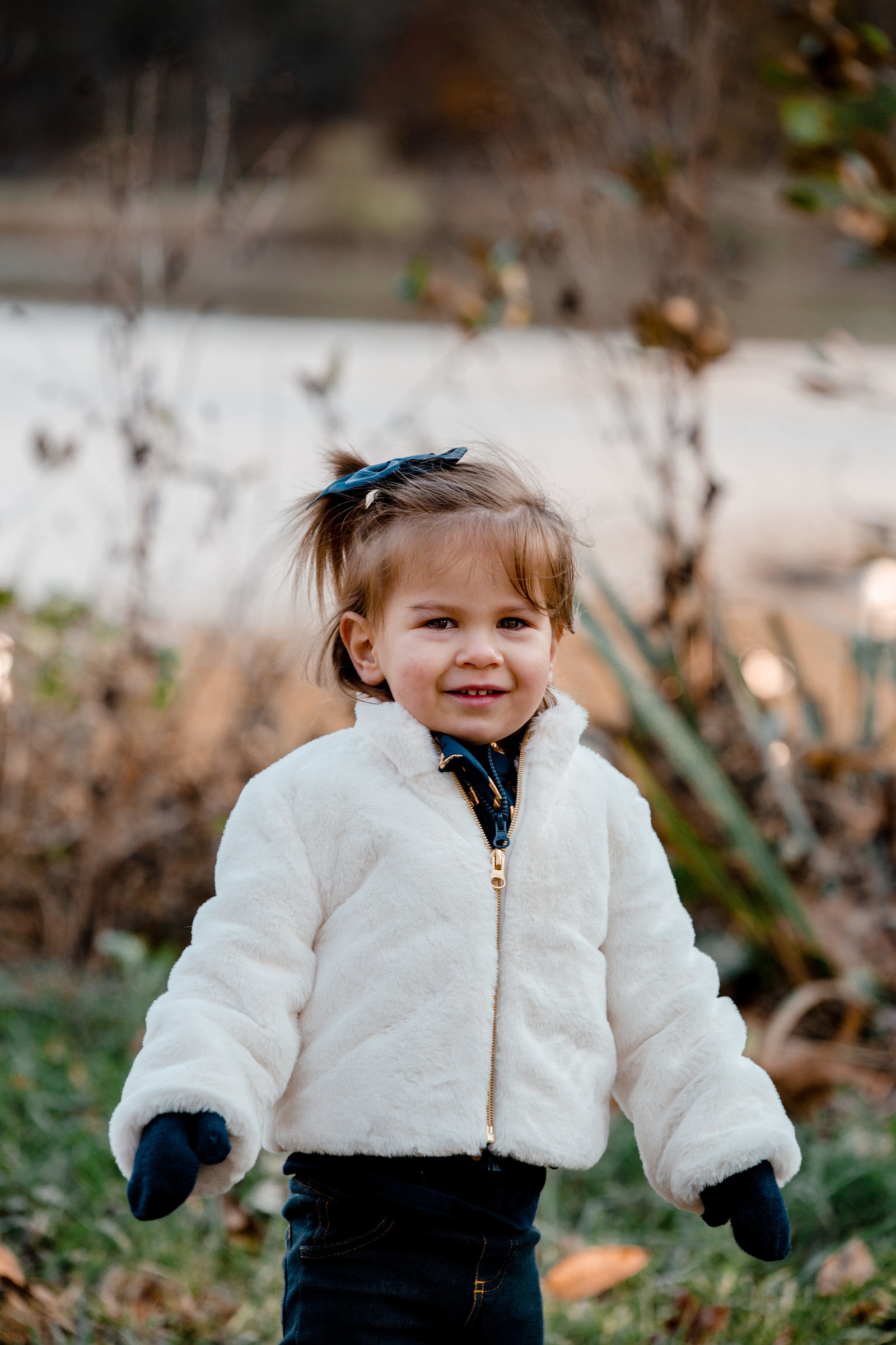 toddler in white fur coat stands in front of pond in north park in the fall