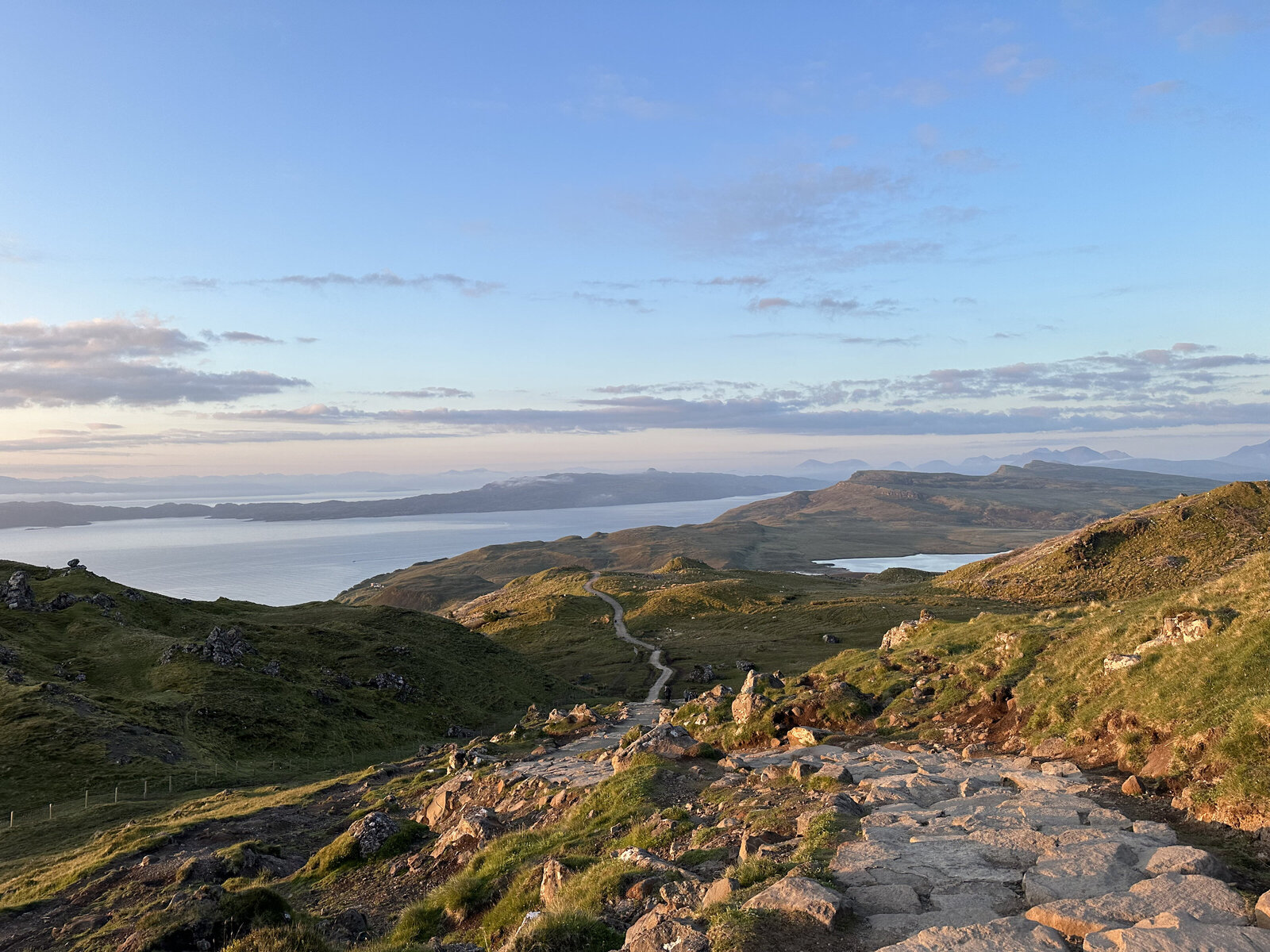 A rocky path winds through green hills toward a distant lake under a clear blue sky at sunset, capturing the spirit of an Oregon coast photography retreat for wild-hearted artists.