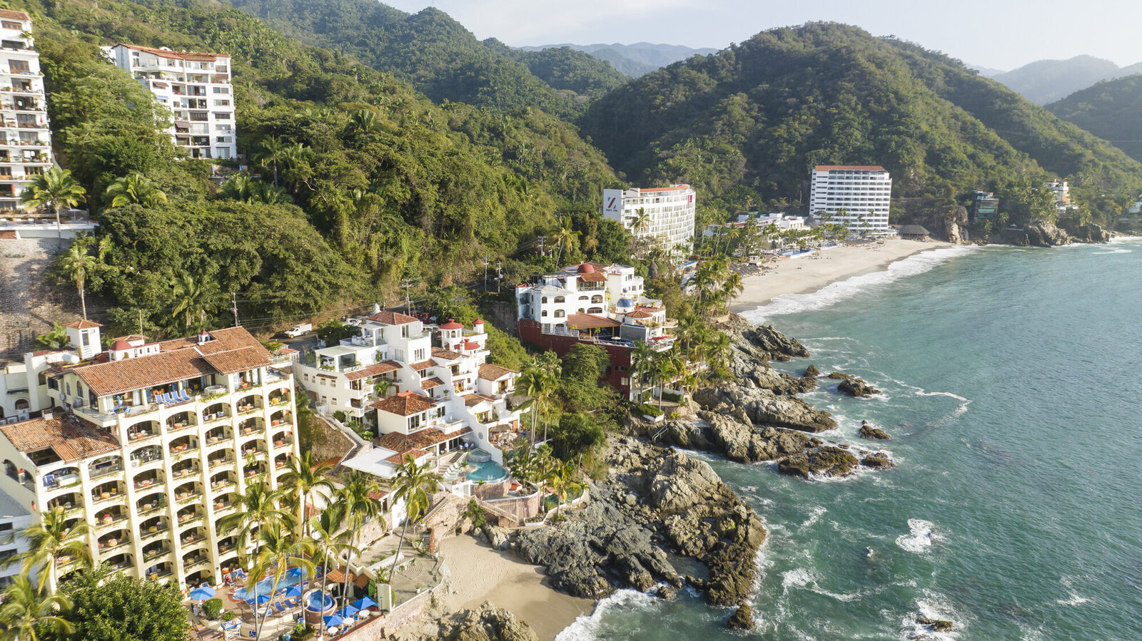 Aerial view of a coastal resort area, a prime location for a destination wedding photographer, with buildings nestled between a lush hillside and a sandy beach.