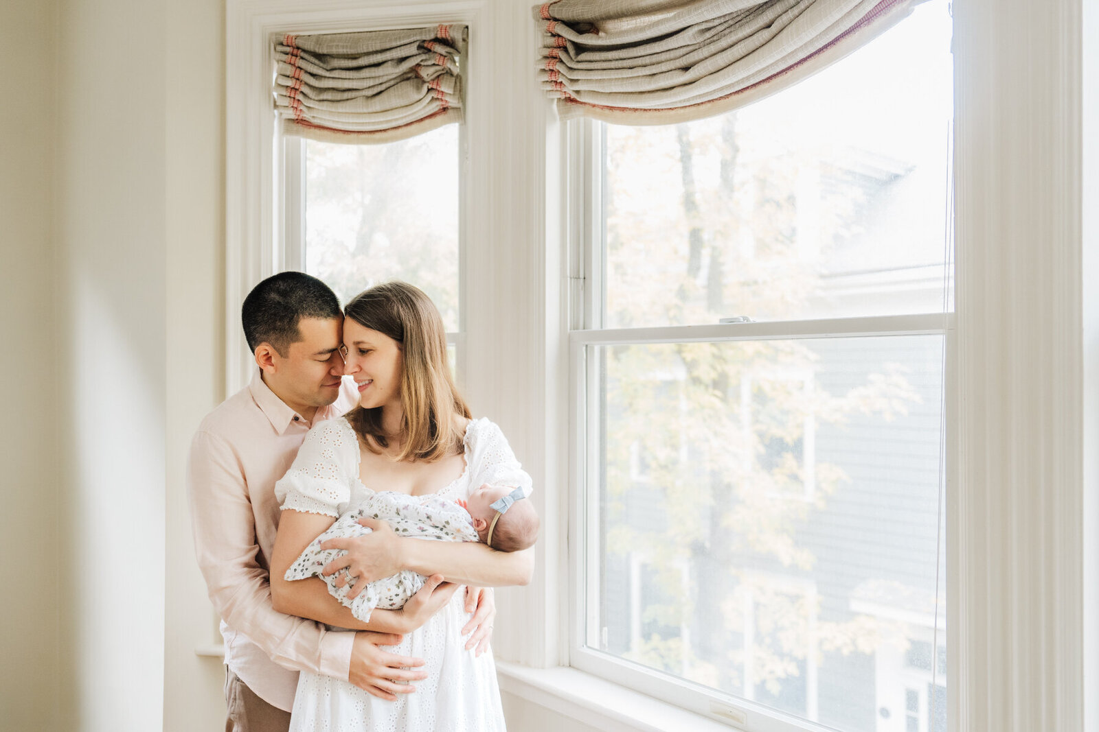new parents stand nose to nose holding baby girl by bay window