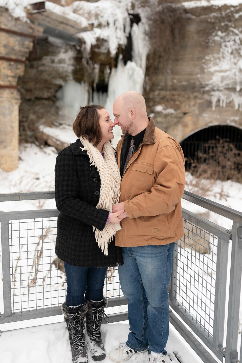 Man and woman in jeans and jackets smile in front of frozen waterfall in Minneapolis, Minnesota.
