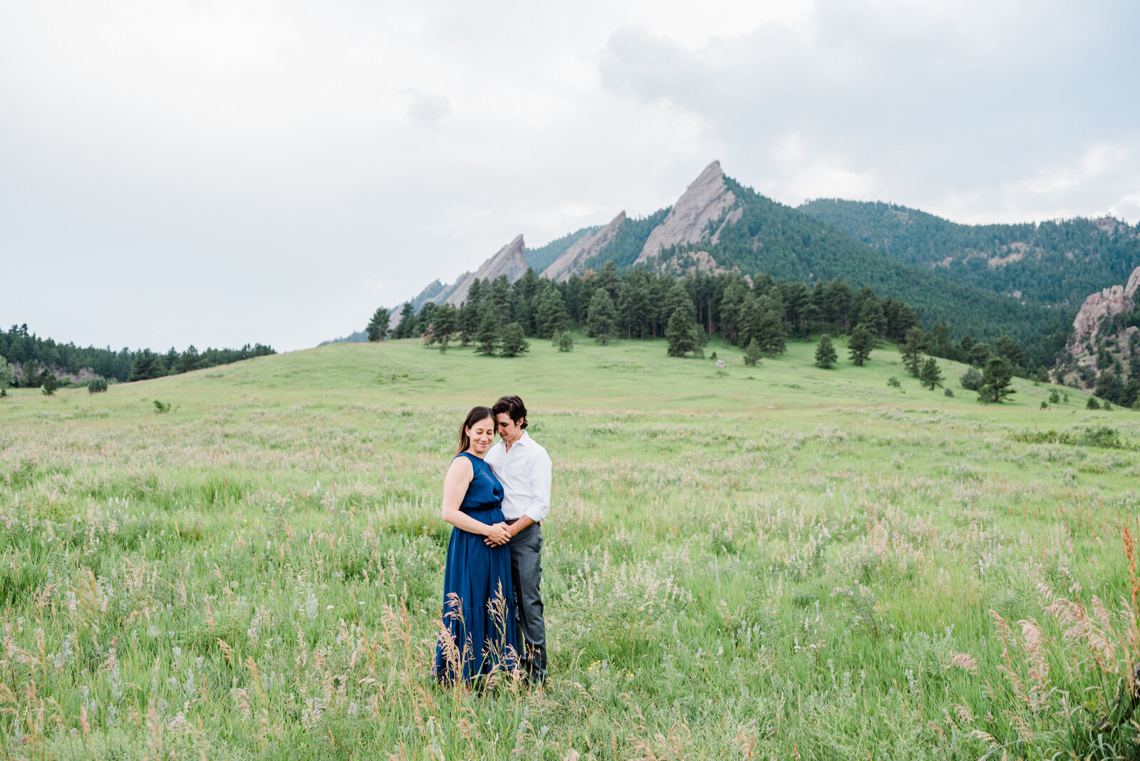 denver family photographers captures maternity photos outdoor with man and woman holding hands as they stand in a field with the Rocky mountains in the distance