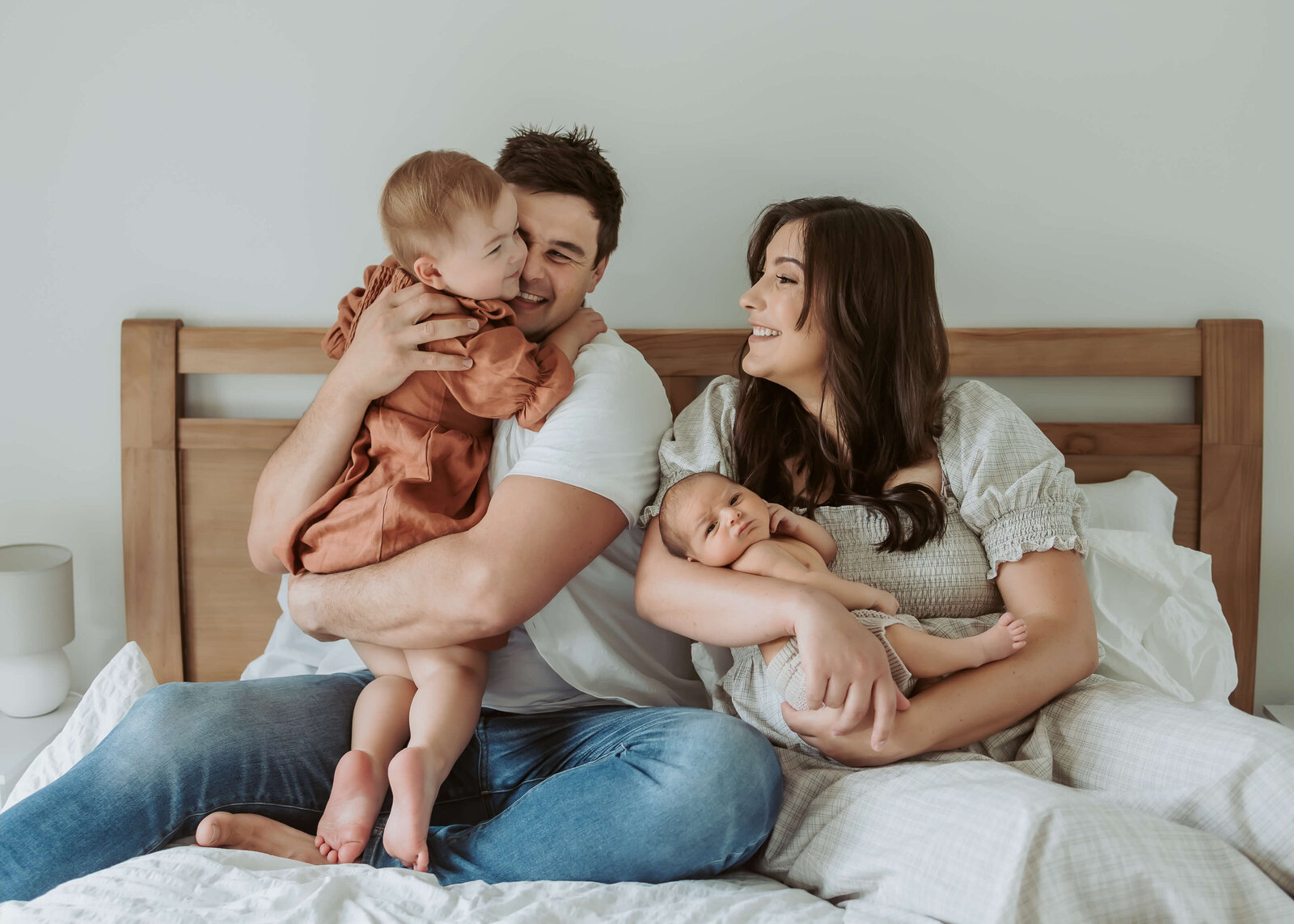 Everyone is on the bed and dad is holding his daughter who is standing with her arms around her neck and mum is looking at them with love while cradling her new baby son in her arms.