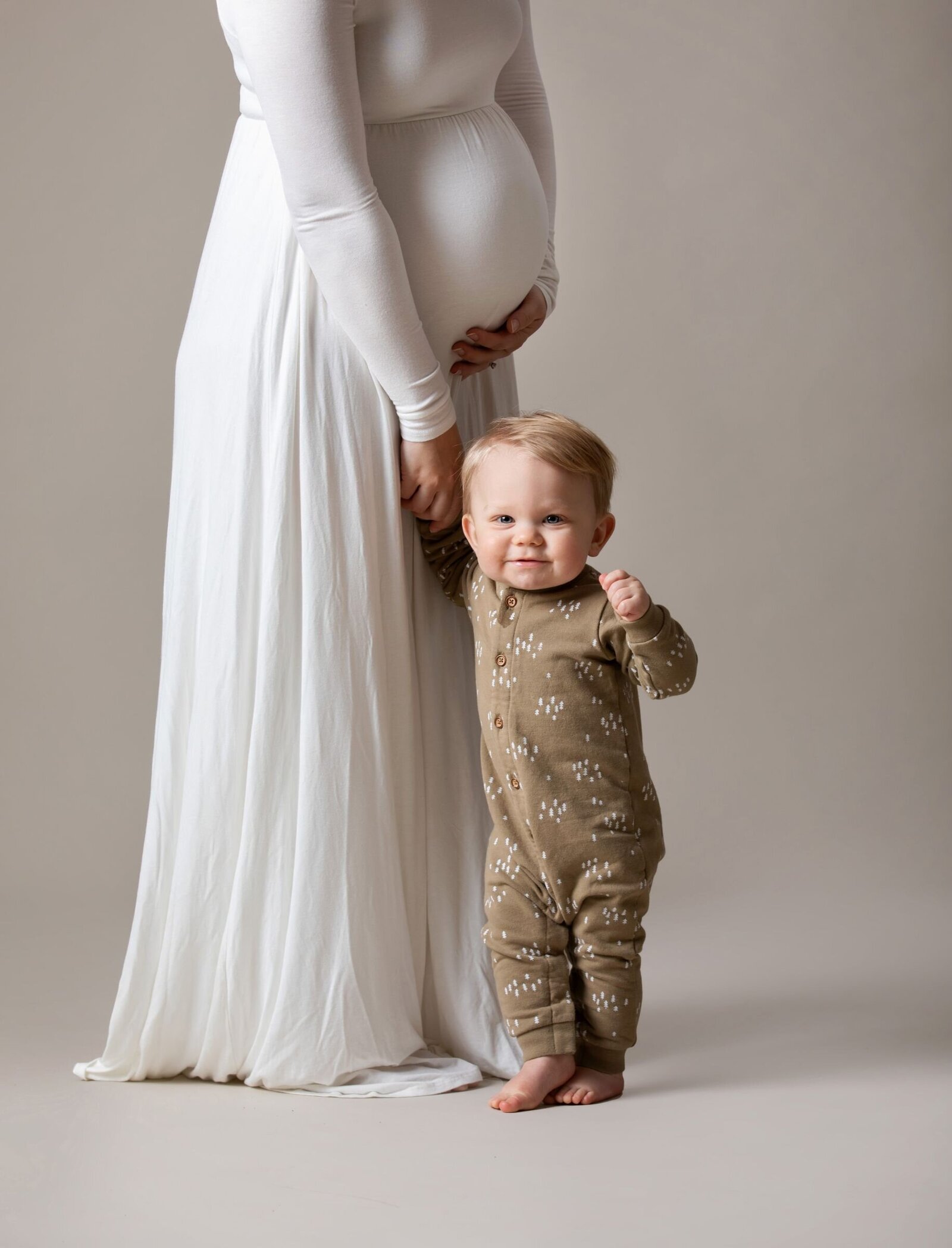 A young mother holding her pregnant belly holding her one year old son's hand standing on a beige backdrop at Studio 64 Photography.
