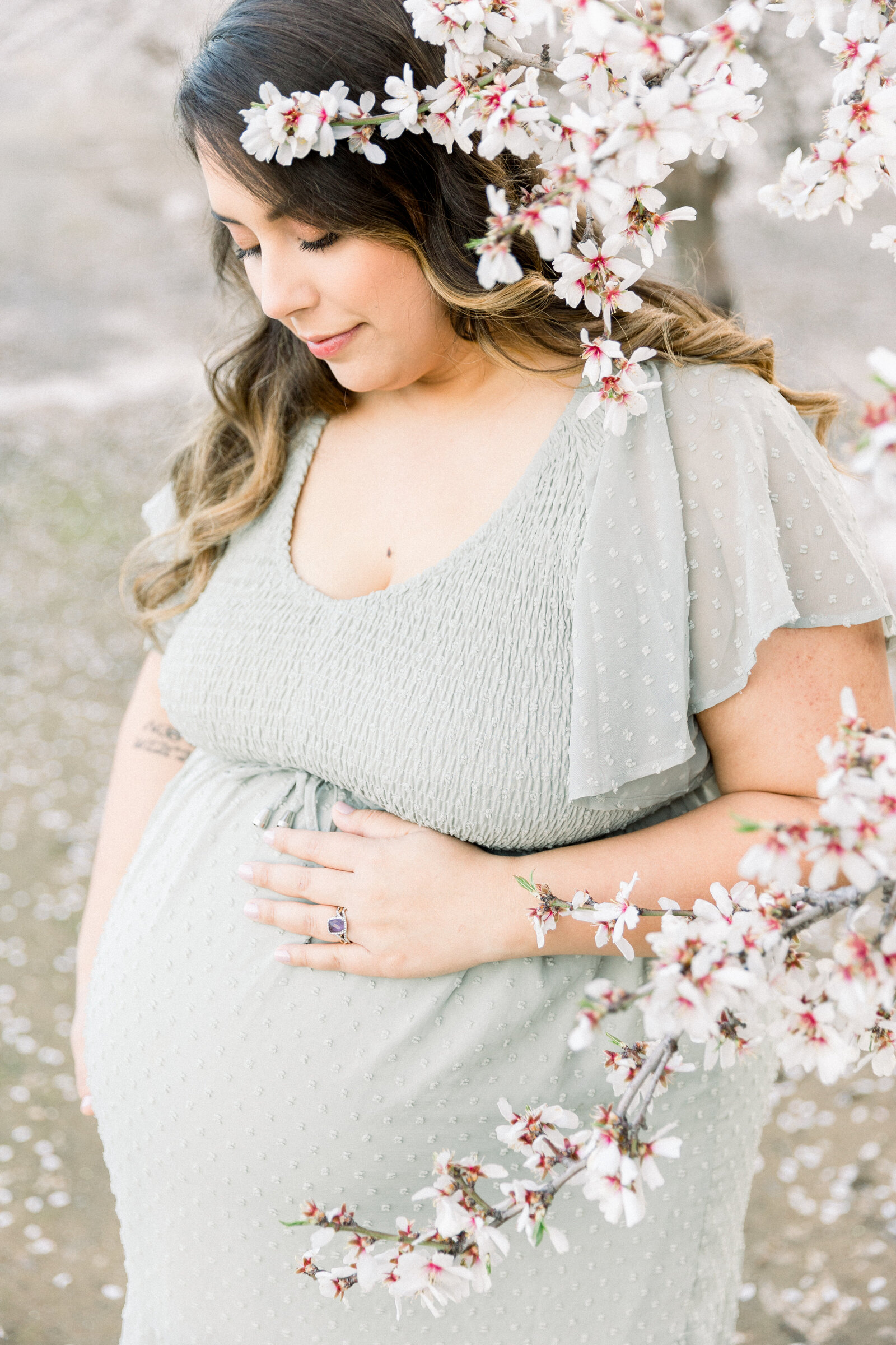 Image of expecting mother looking down at baby bump standing in almond orchard taken by Sacramento Newborn Photographer Kelsey Krall