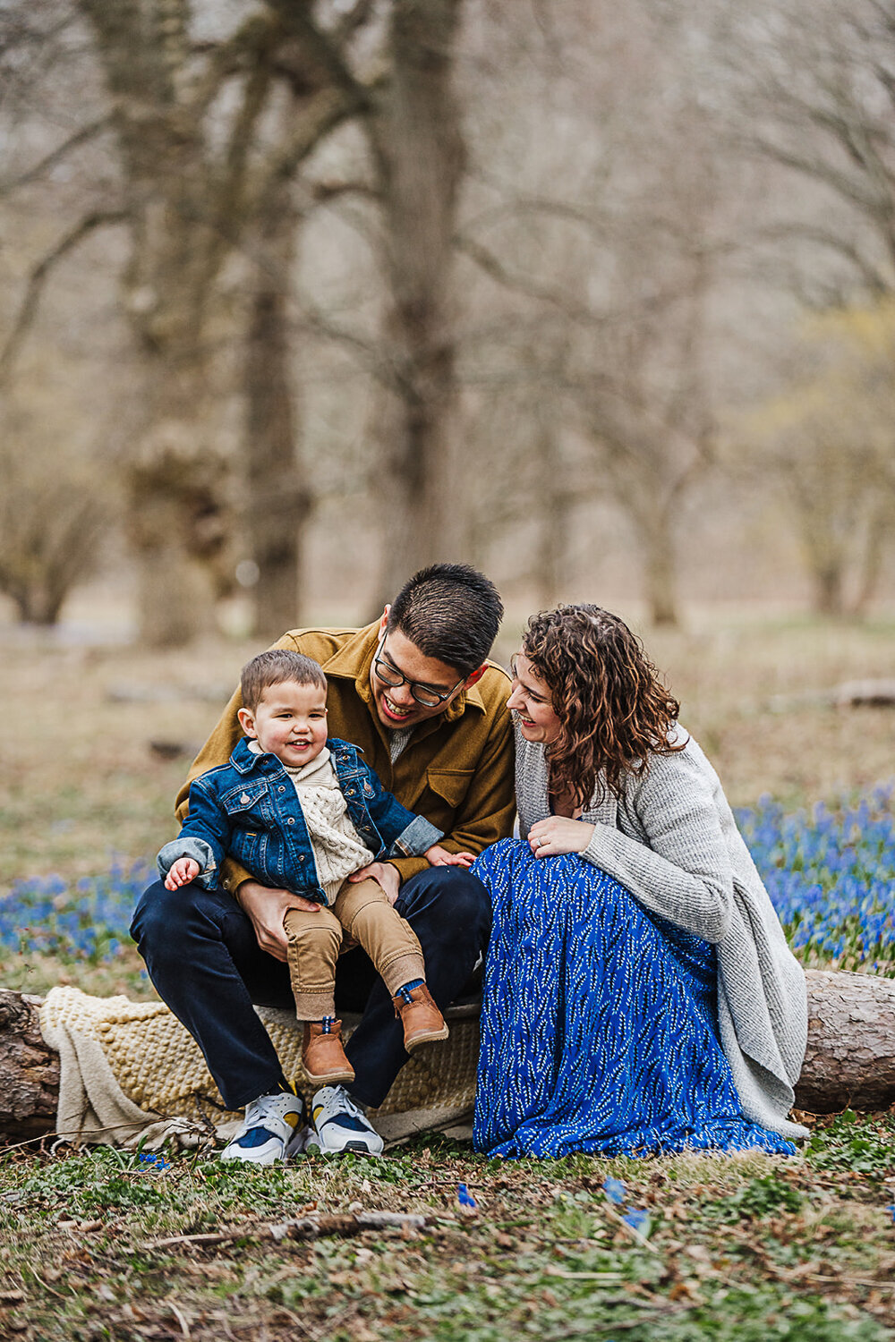 couple sit on log with toddler boy among blue ground flowers
