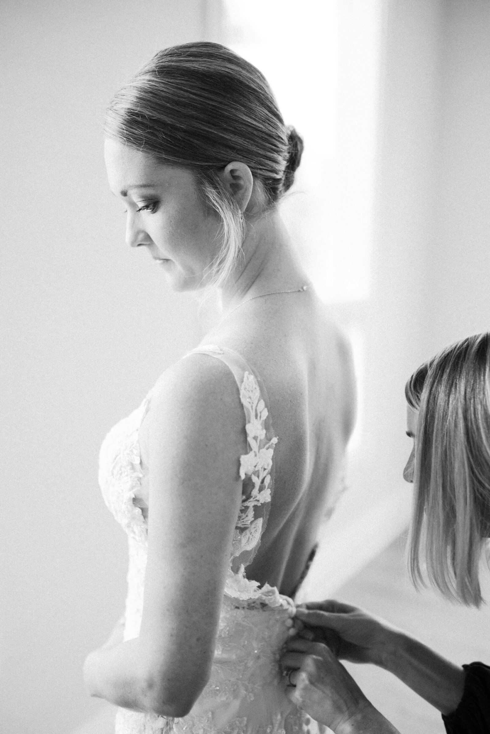 black and white photograph of bride  looking down toward the floor while her maid of honor helps to close the back of her dress