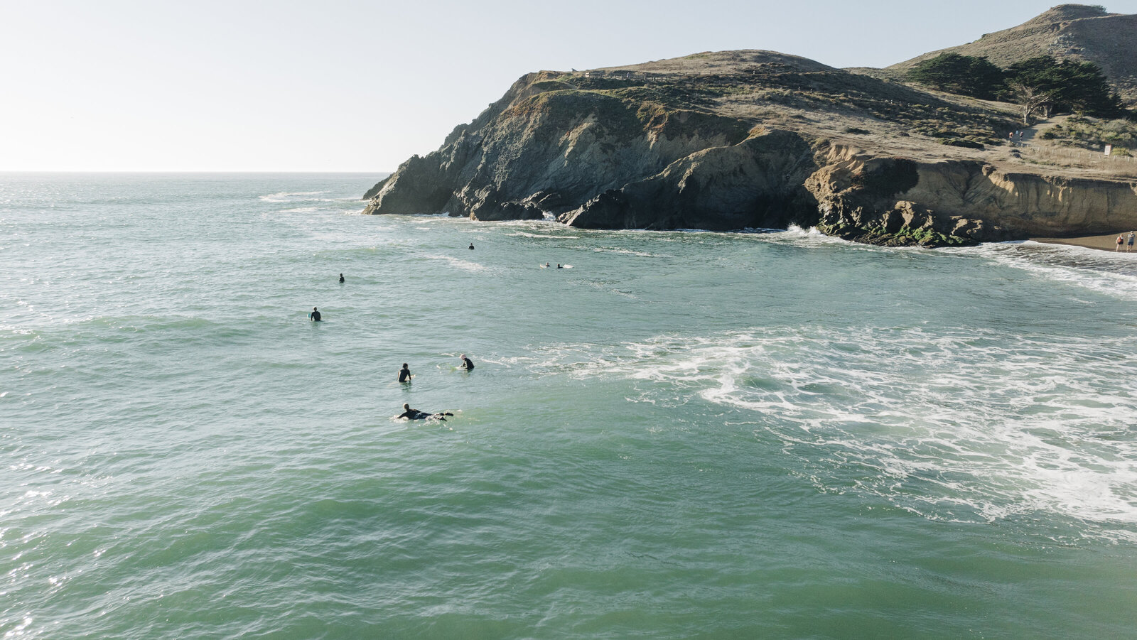 Surfers in the ocean near a rugged cliff under a clear sky, captured by a Destination Wedding Photographer.