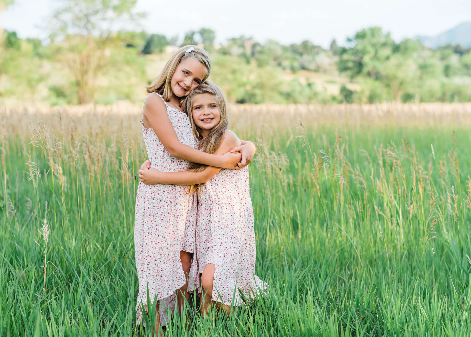 Two young blonde sisters in pink dresses hug each other while standing in a green field in an image by Northern Virginia family photographer
