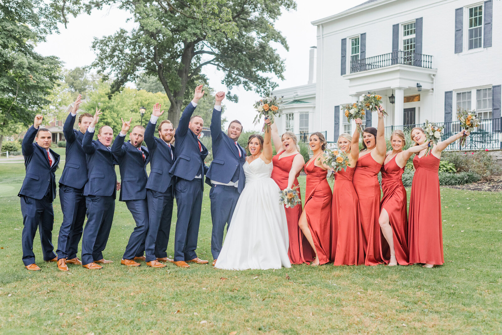 bride and groom stand in the middle of their bridal party raising their hands with joy