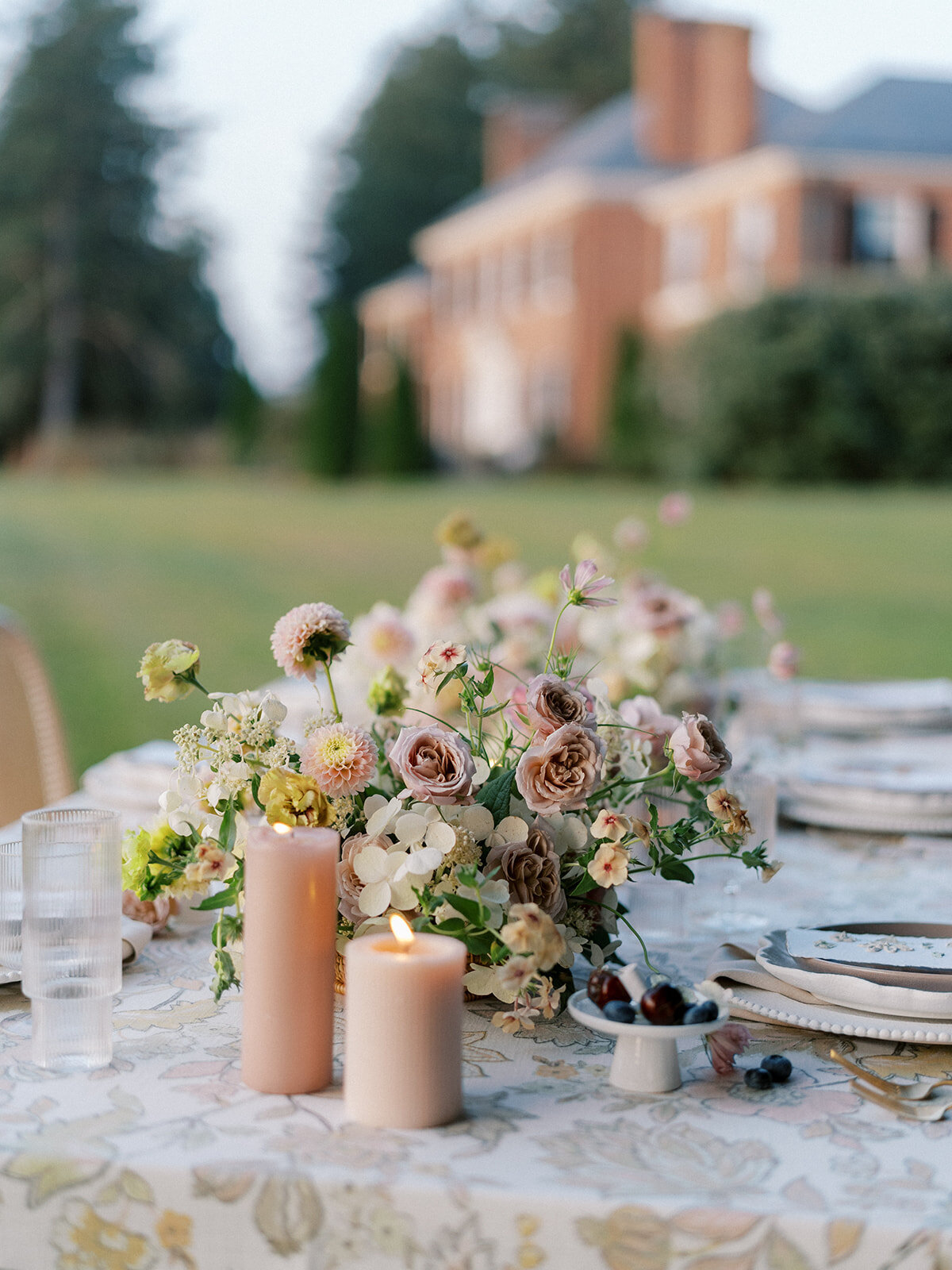 Long table with floral-patterned linen with blush pillar candles, fresh fruit and blush, taupe, cream and mauve wispy floral compotes going down the length of the table.