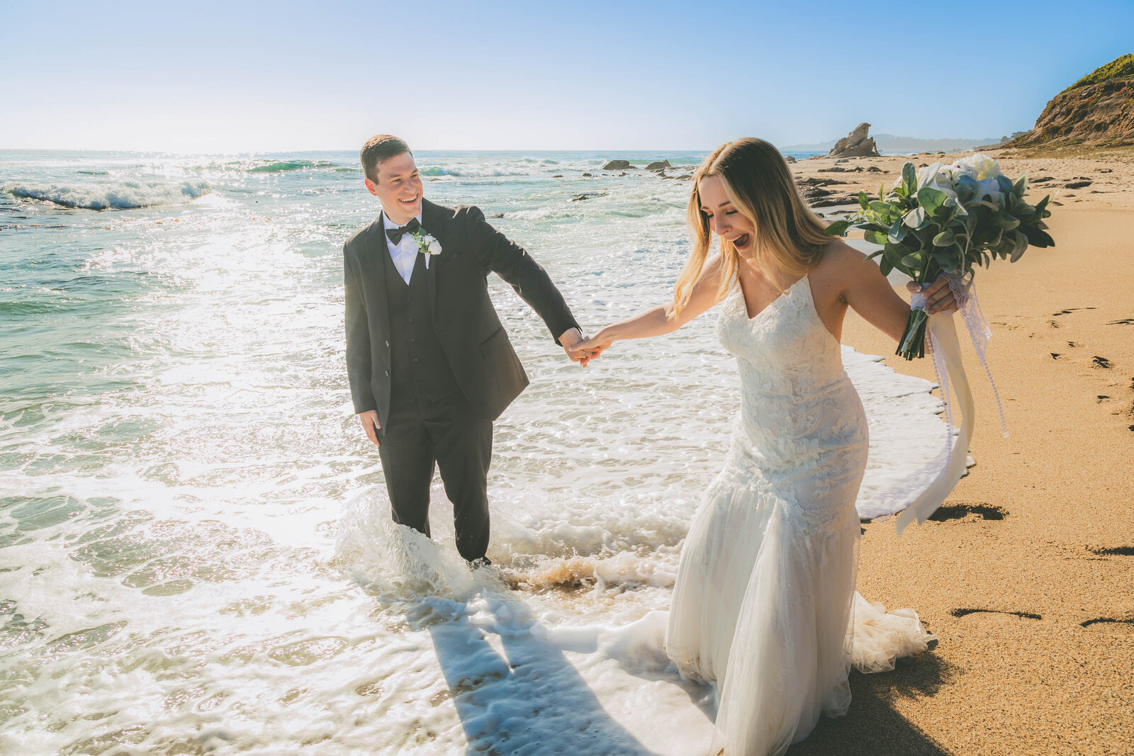 A couple who just got married laugh as they get wet from the waves during their beach wedding.