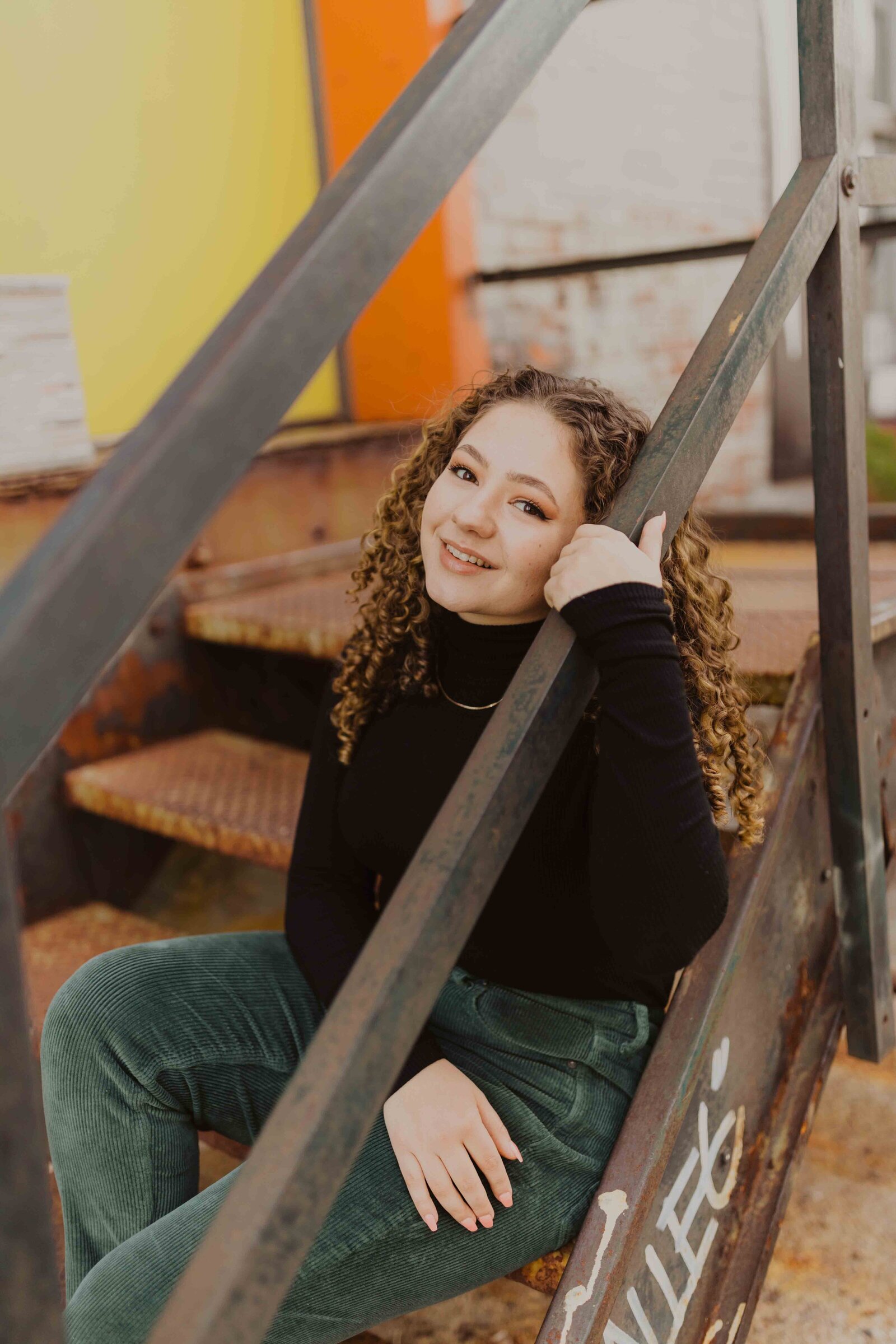 Senior girl sitting on metal stairs, holding onto railing and smiling.