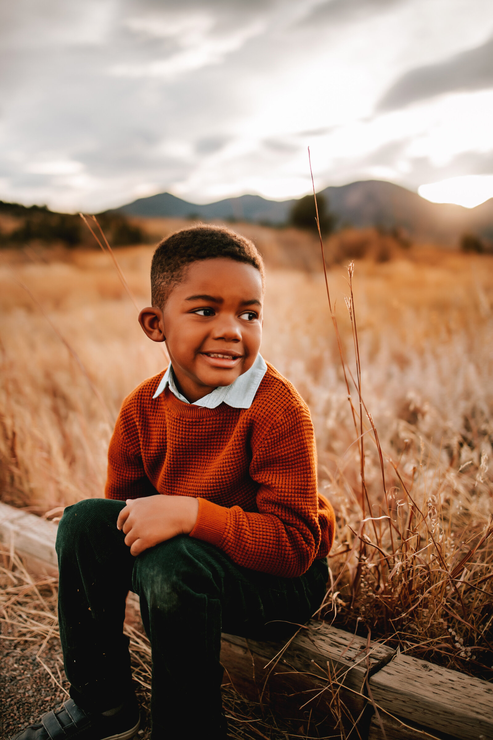 young african american boy in an orange swearter sits on a log with mountains behind him