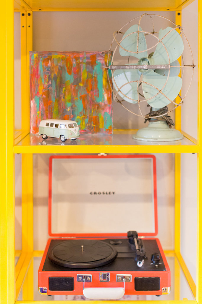 Yellow and glass shelves with an orange record player, fan, art, and mini VW.