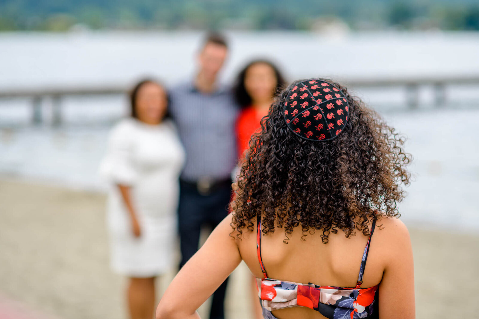 A teen girl in a kippah stands on a beach looking at her parents in the distance during a Bellevue Bar and Bat Mitzvah Photography session