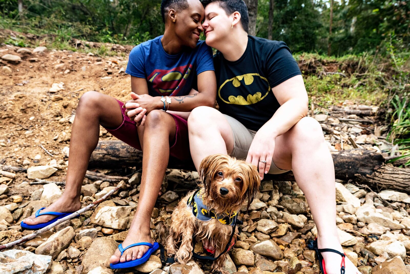 same-sex couple in superhero shirts sits on the river bank at the Eno River