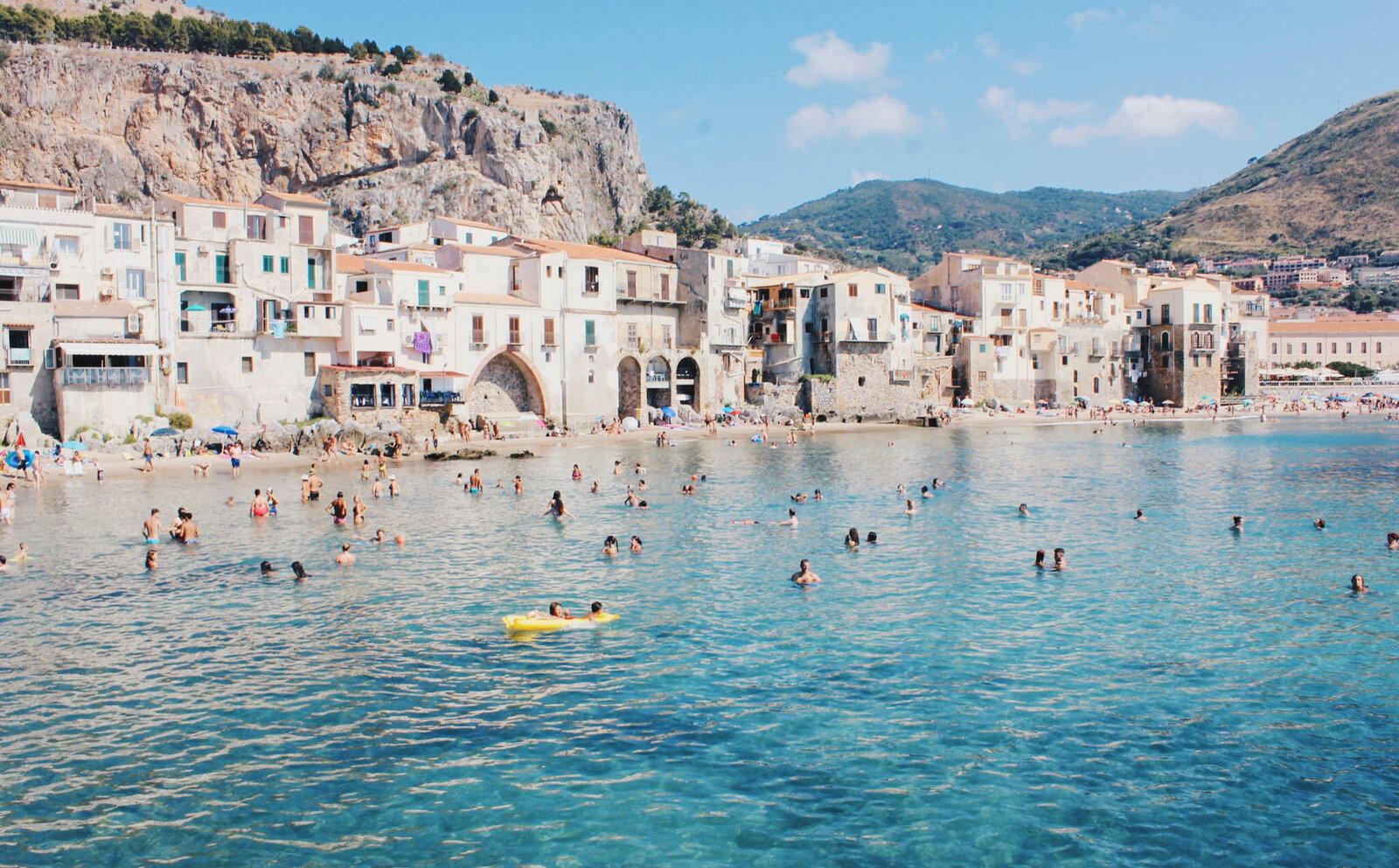 A scenic beach in Cefalù, Italy, features clear blue water with many people swimming and relaxing. Historic buildings and cliffs line the shore, while distant mountains provide a picturesque backdrop under a sunny sky.