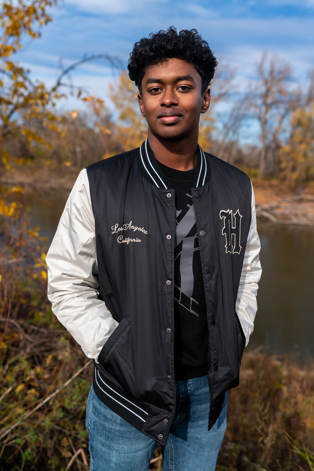 Senior Indian boy smiles in letter jacket in front of the lake.