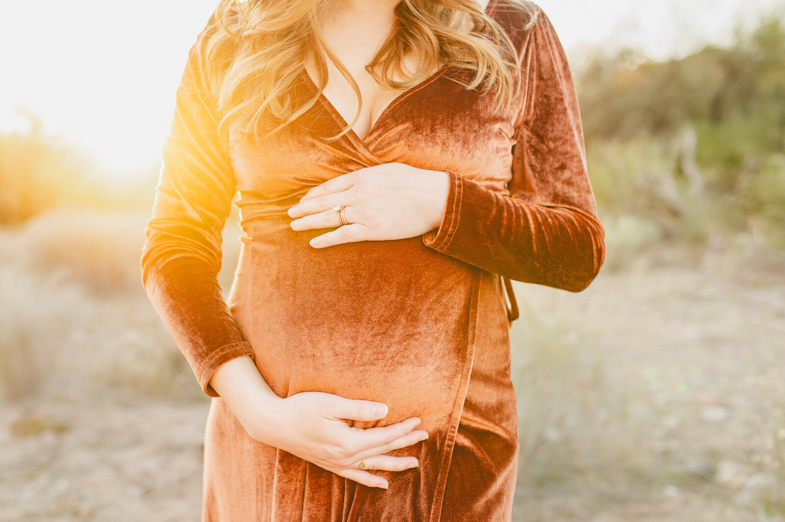 pregnant mom holding her tummy glowing among the Phoenix sunset in a burnt orange velvet maternity gown