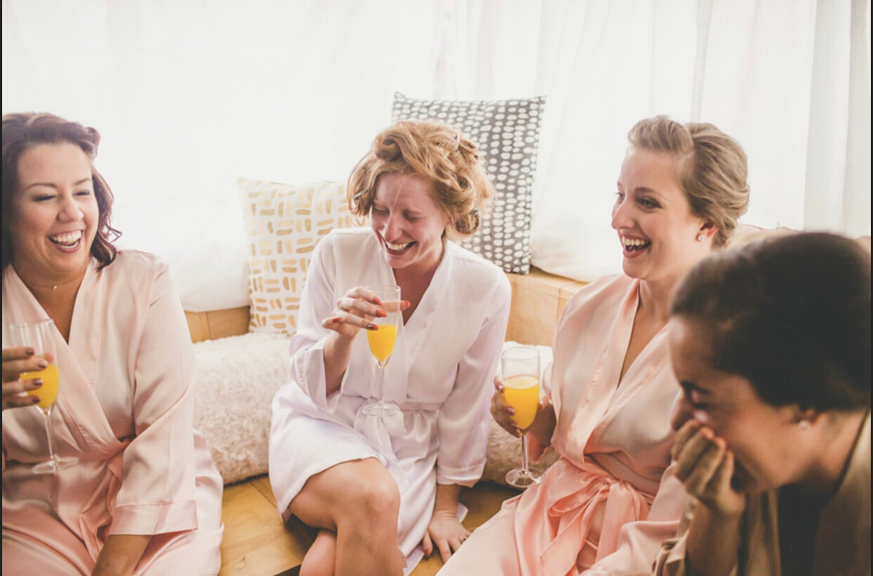 a bride and her 3 bridesmaids, wearing robes, drinking mimosas while laughing and getting ready for the wedding.