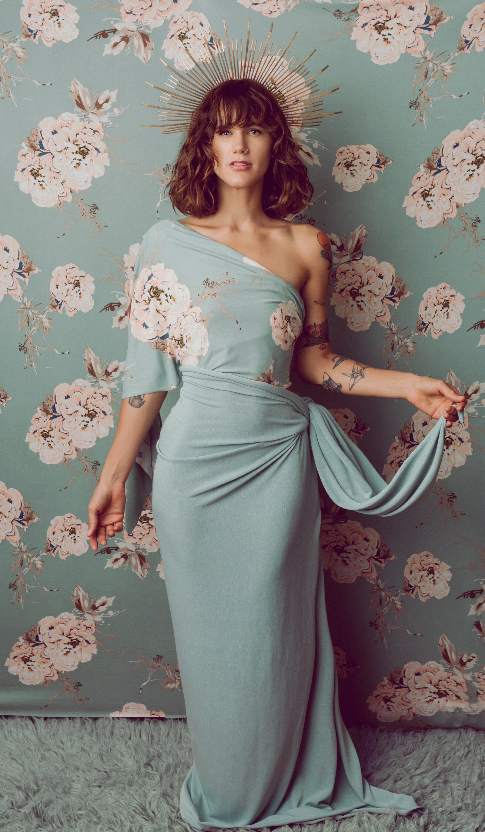 Portrait of a woman in a light blue gown in front of wallpaper that matches the gown, facing camera center.