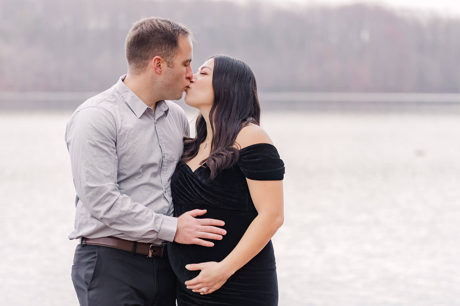 An expecting couple kissing and embracing mom’s belly in front of Burke Lake.