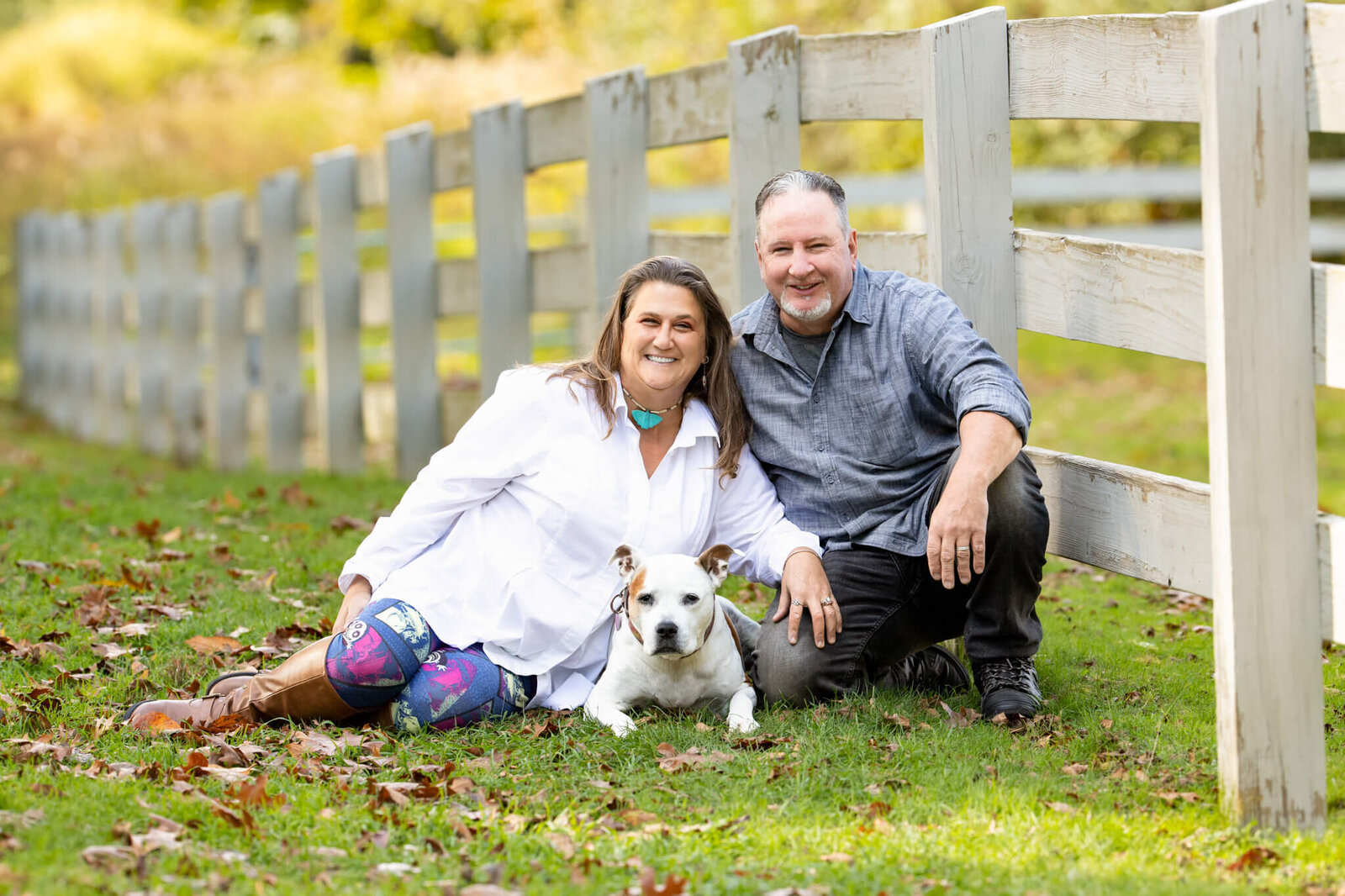 A couple and their dog sitting at a white barn fence with leaves on the ground in fall snuggling with their white dog