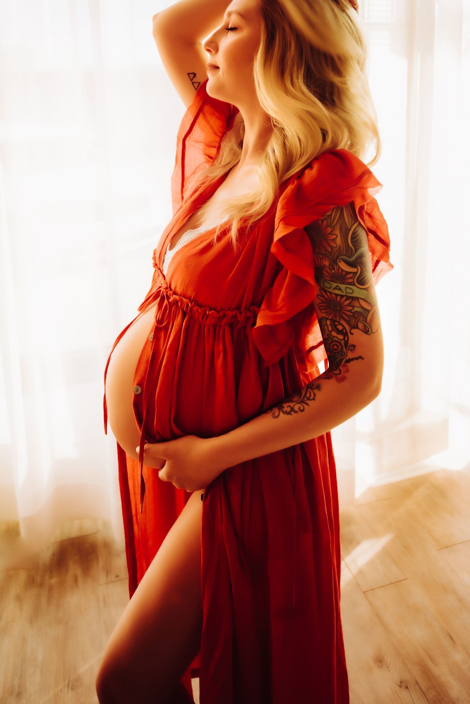 a pregnant woman in a red dress that exposes her belly. she is standing with one arm over her head and the other hand gently holding her belly. captured by Infinite Productions