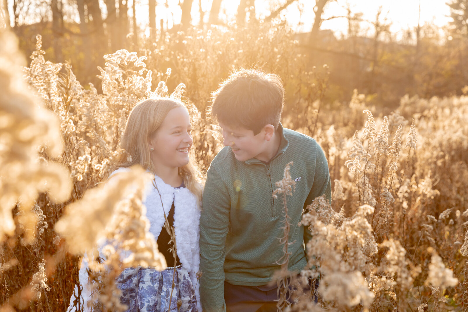 brother and sister stand in field of tall grass during golden hour while looking at each other smiling and laughing