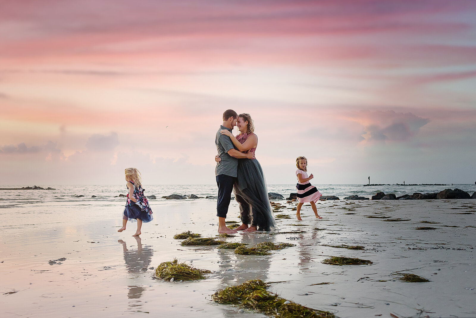 Mom and dad embracing while daughters run around them on beach in Florida.