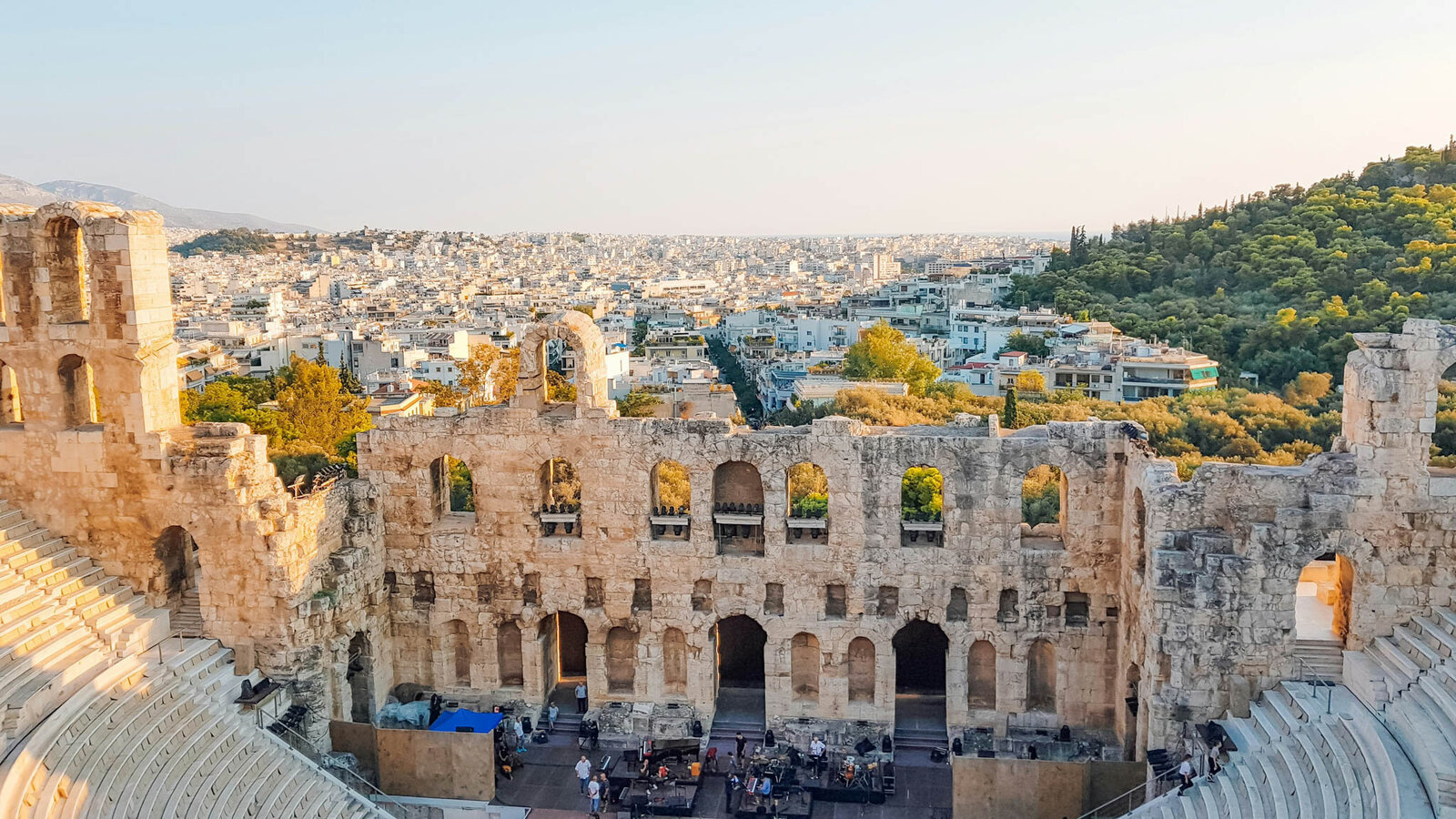 The image shows the ancient Odeon of Herodes Atticus amphitheater in Athens, Greece. The stone structure is partially intact, with scenic views of the city and lush greenery in the background under a clear sky.