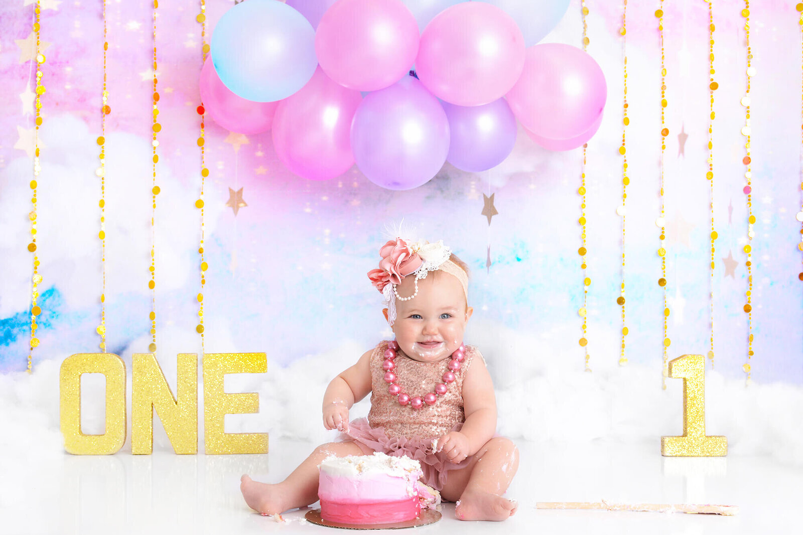 girl smiles at the camera with a pink and white birthday cake with pink blue and purple balloons above her