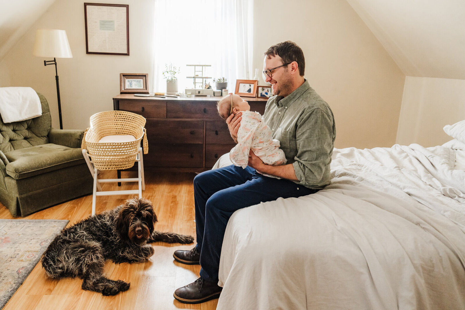 dad sits on master bed holding baby girl while brown dog looks on