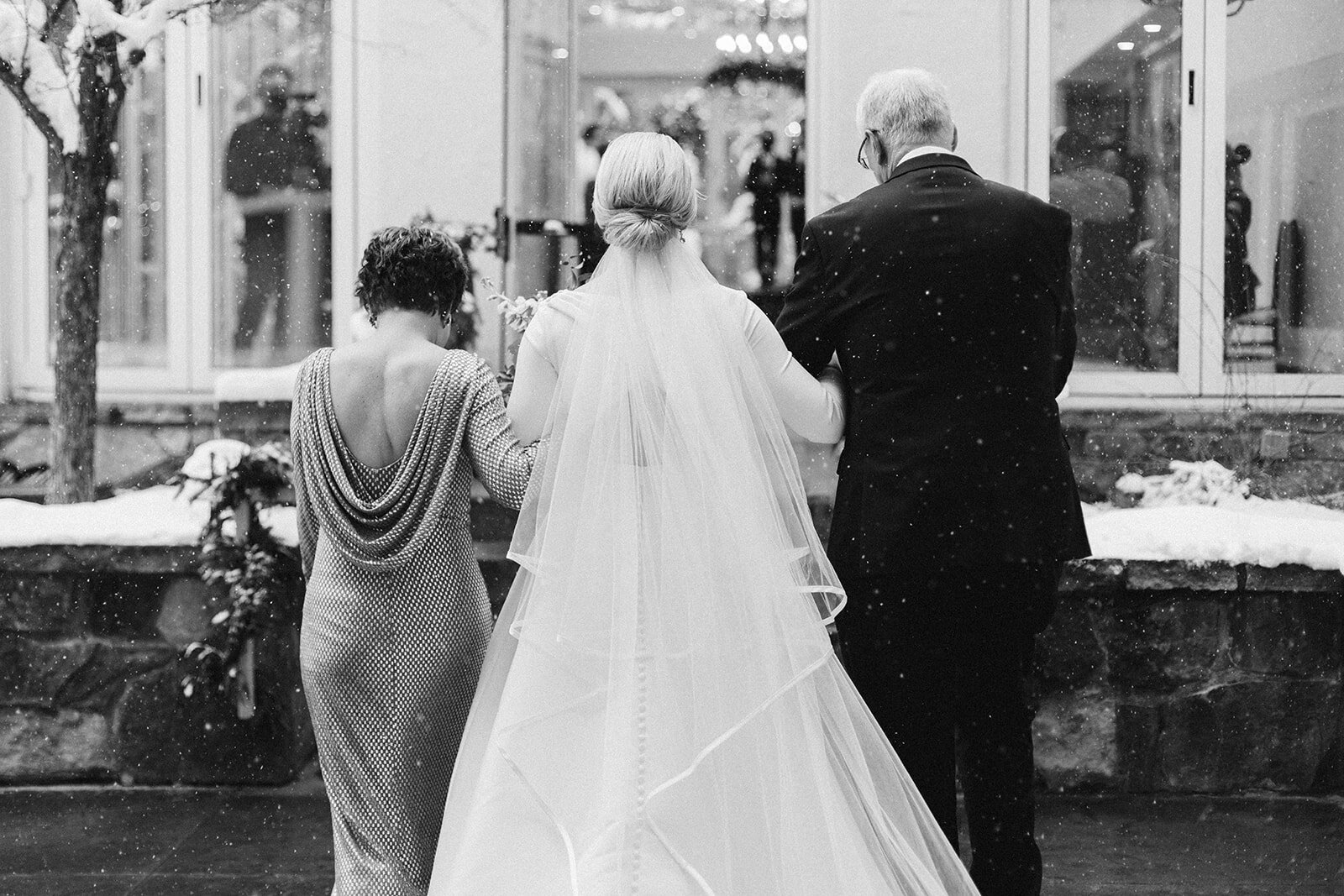 Parents walk bride down the aisle while its snowing in black and white