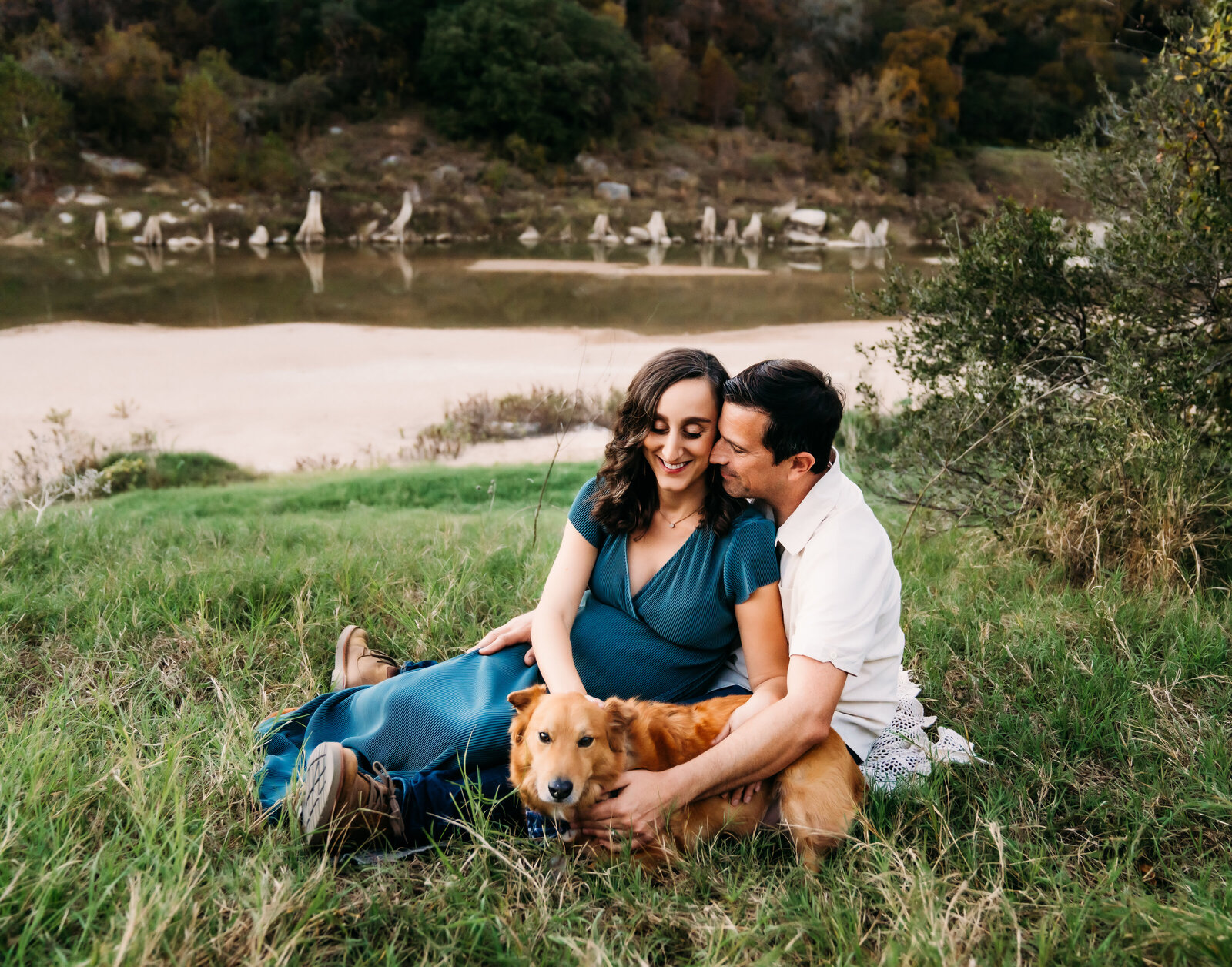Maternity Photographer, man and pregnant wife sit in grass beside the lake with their dog