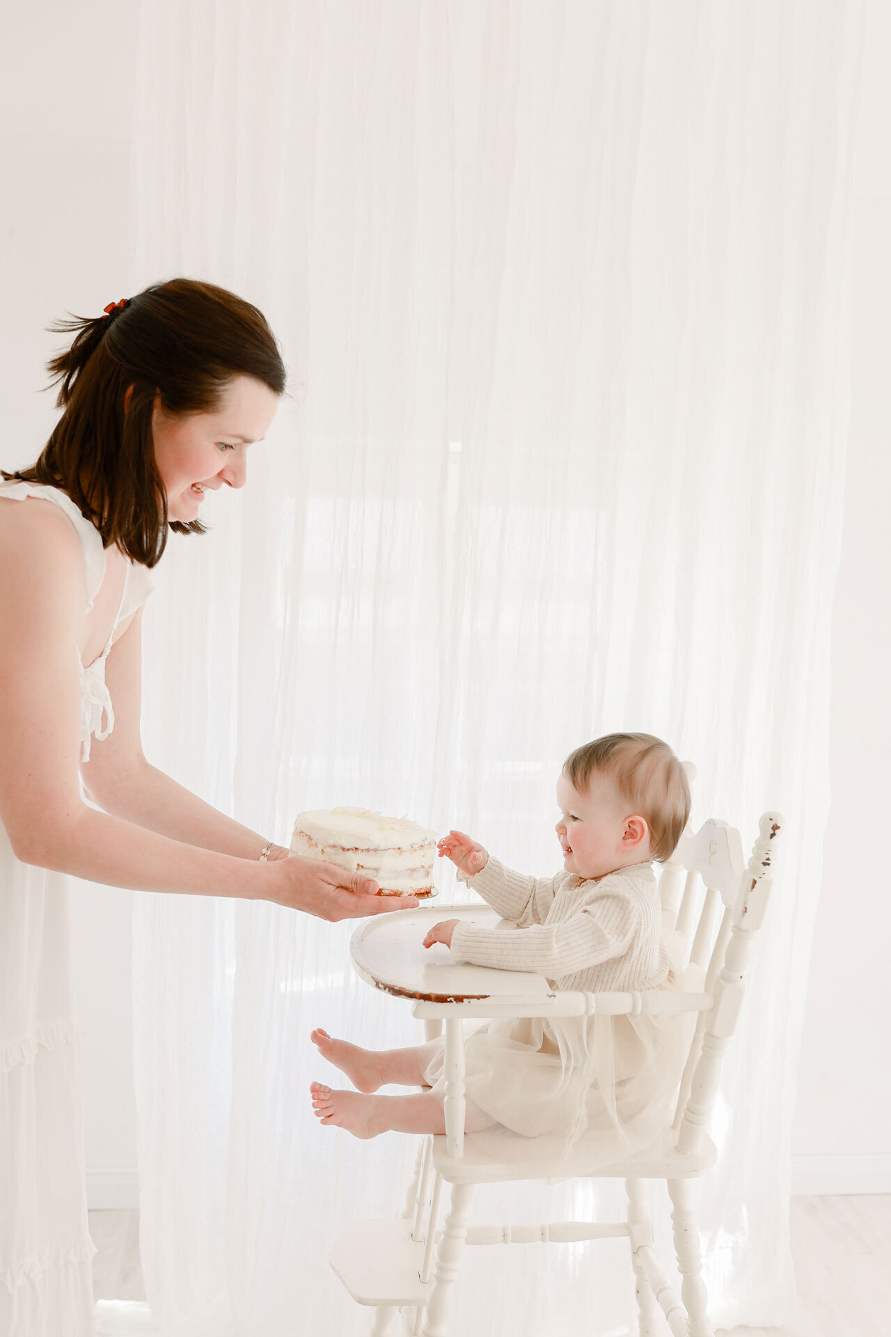 Baby Fotoshooting von Babys erstem Geburtstag. Fröhliches Baby in einem Hochstuhl freut sich auf den Kuchen den Mama ihm reicht.